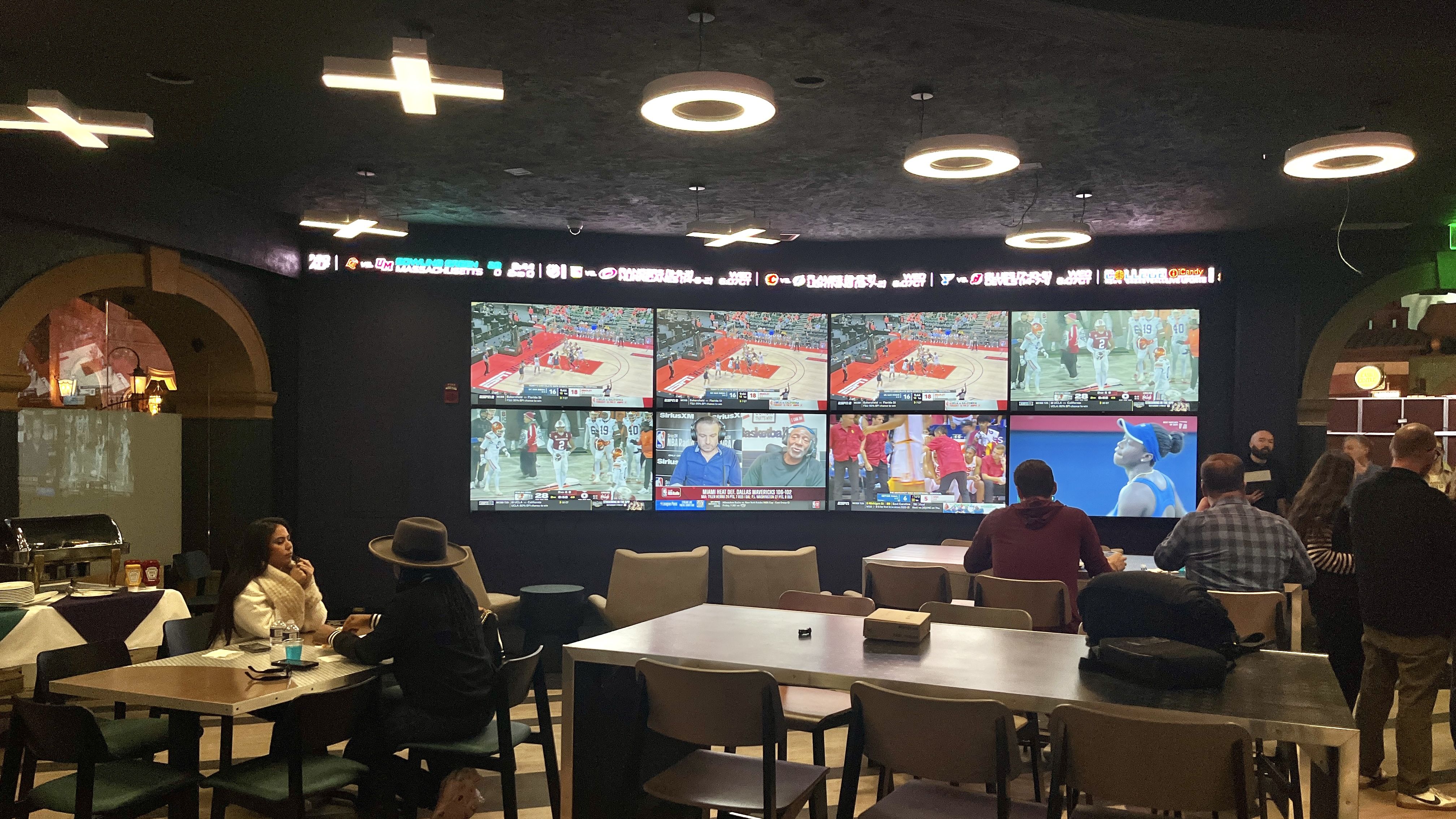 A dimly lit sports bar with multiple large screens showing different sports broadcasts, including basketball and football. Several people are seated at tables watching the games.