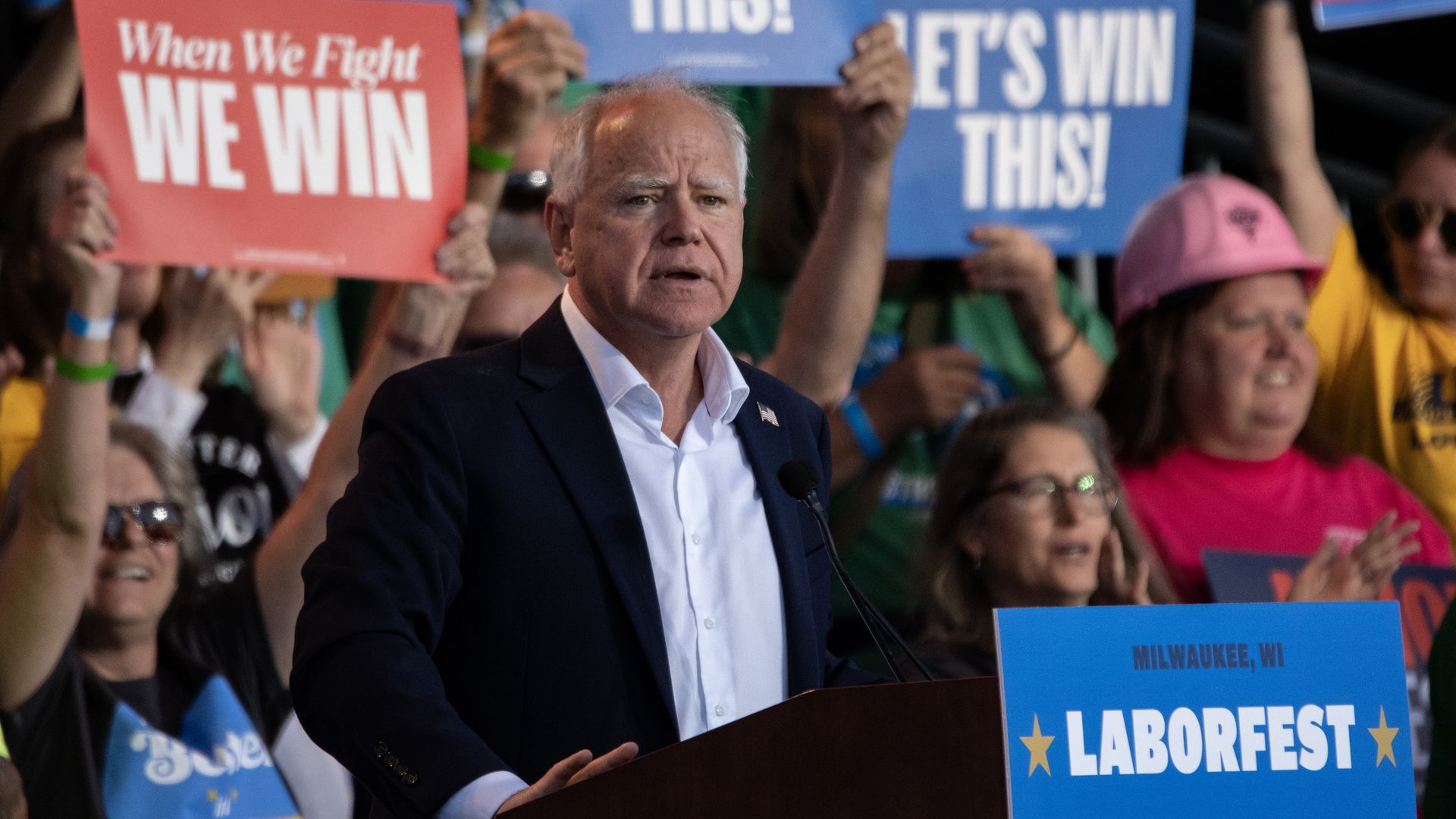 Minnesota Gov. Tim Walz, wearing a tieless blue suit, standing at a podium with a blue "LABORFEST" sign in front of supporters holding blue and red "LET'S WIN THIS" and "When We Fight WE WIN" signs.