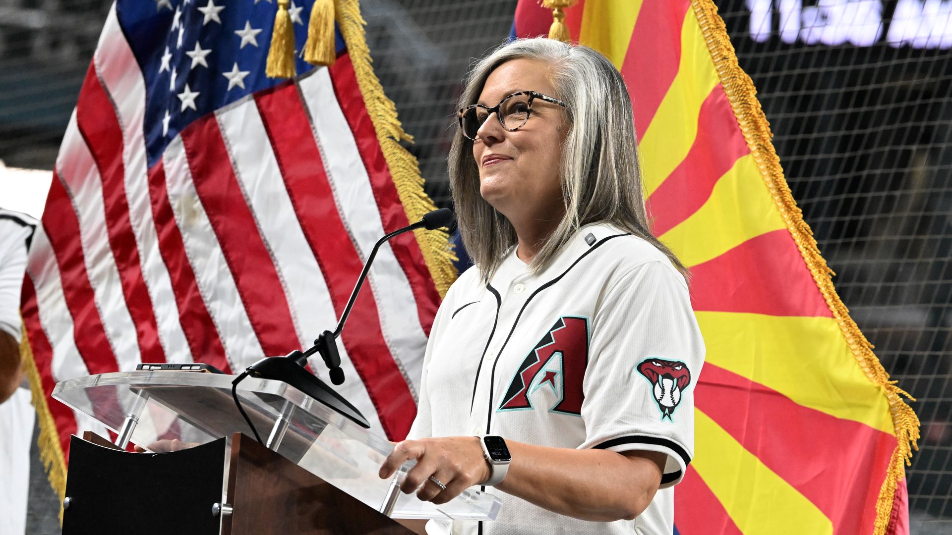 A smiling woman in a white baseball jersey with a red A on the front and a red snake head with a baseball in its mouth on the sleeve. The woman is standing at a lectern with a microphone, with U.S. and Arizona flags behind her. 