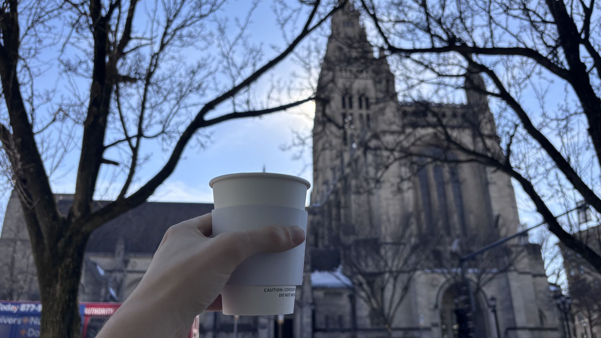 Hand holding a white paper cup with a sleeve in front of a tall old stone cathedral with leafless tree branches framing the scene under a clear blue sky.