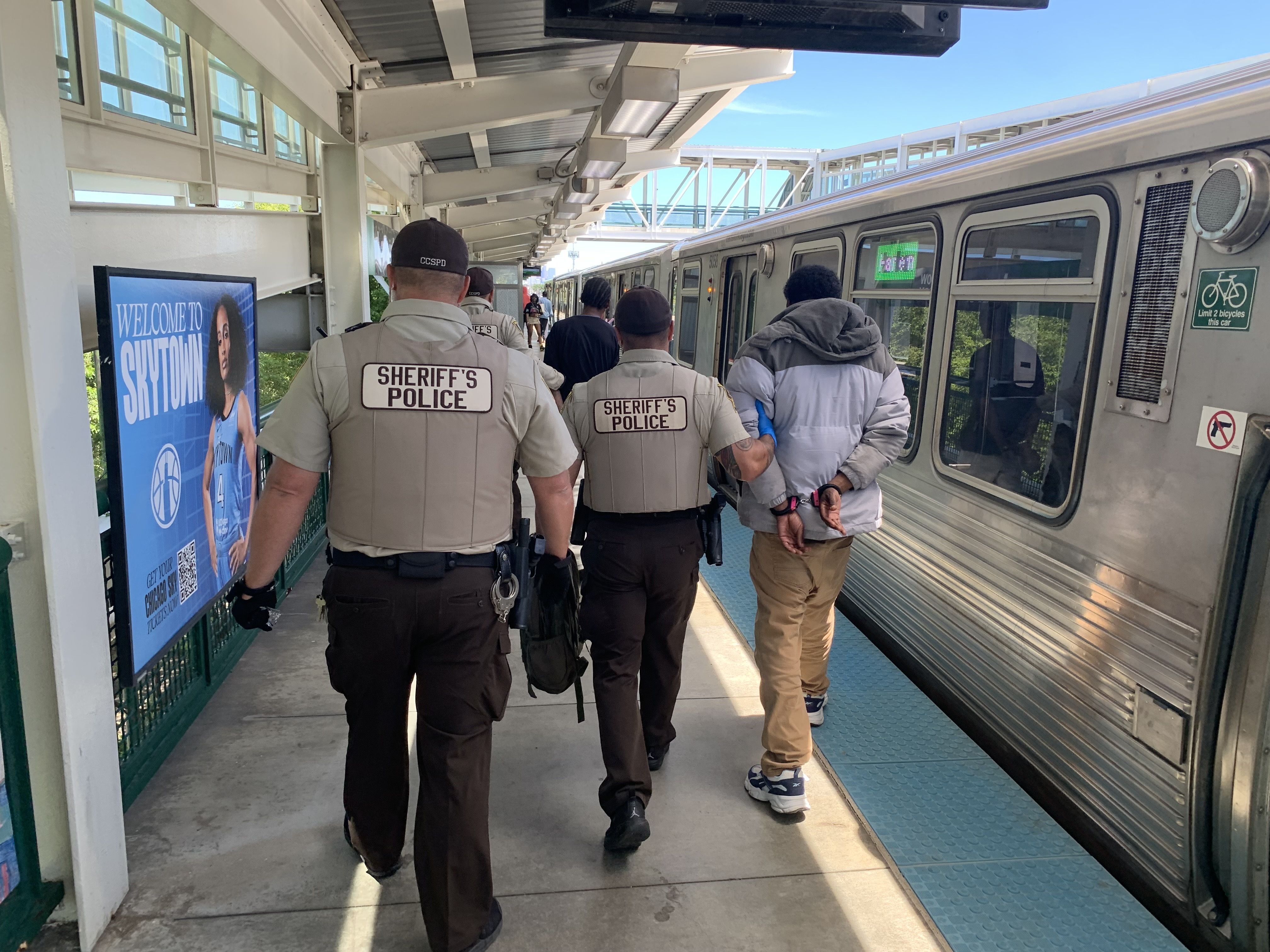 Back view of Sheriff’s Police officers in tan uniforms guiding passengers along an elevated platform beside a silver subway car; blue poster, bicycle sign, and bright sunny day.