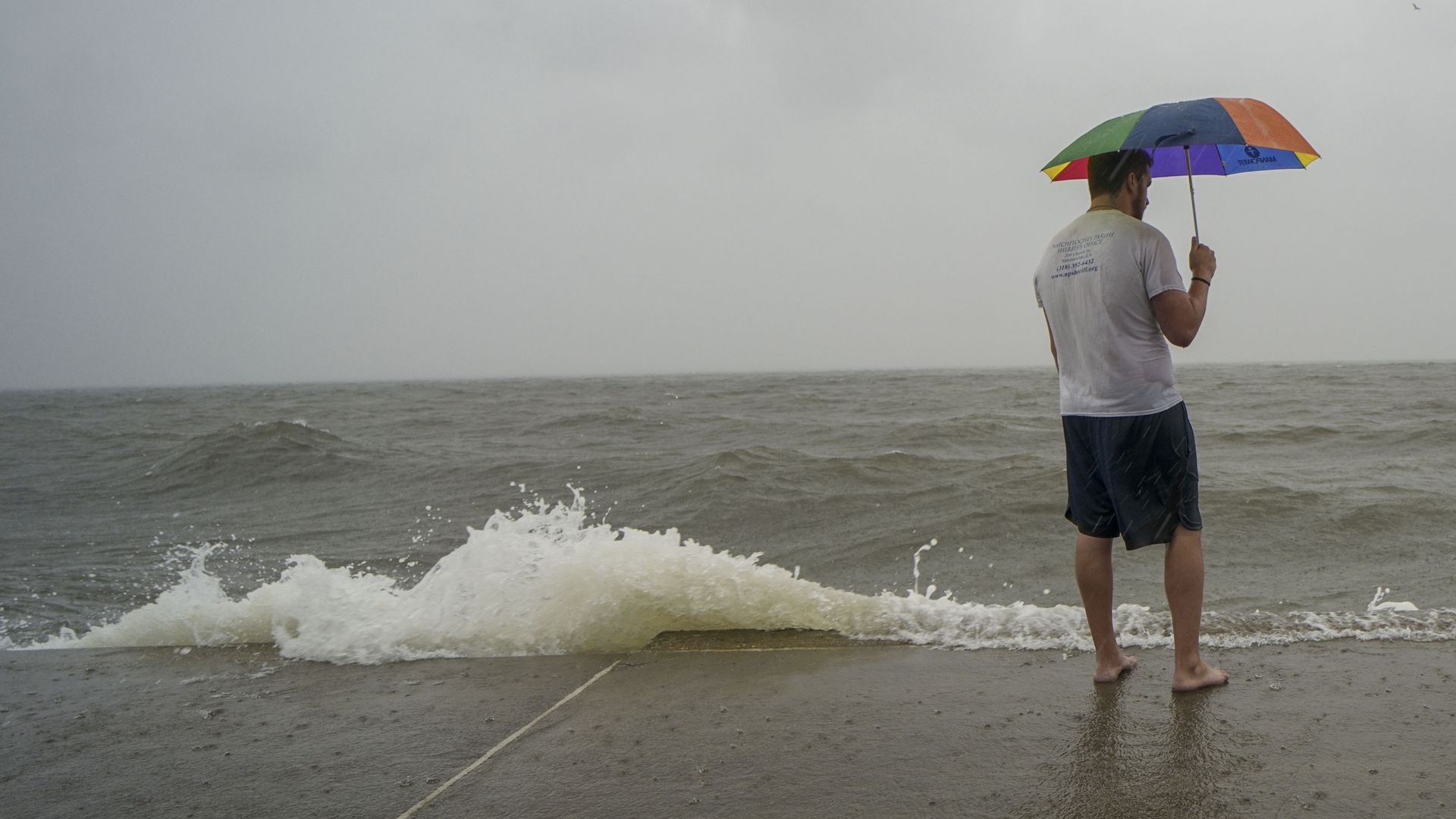 A man stands barefoot next to Lake Pontchartrain as waves crest. He is wearing a T-shirt and shorts while holding a rainbow-colored umbrella.