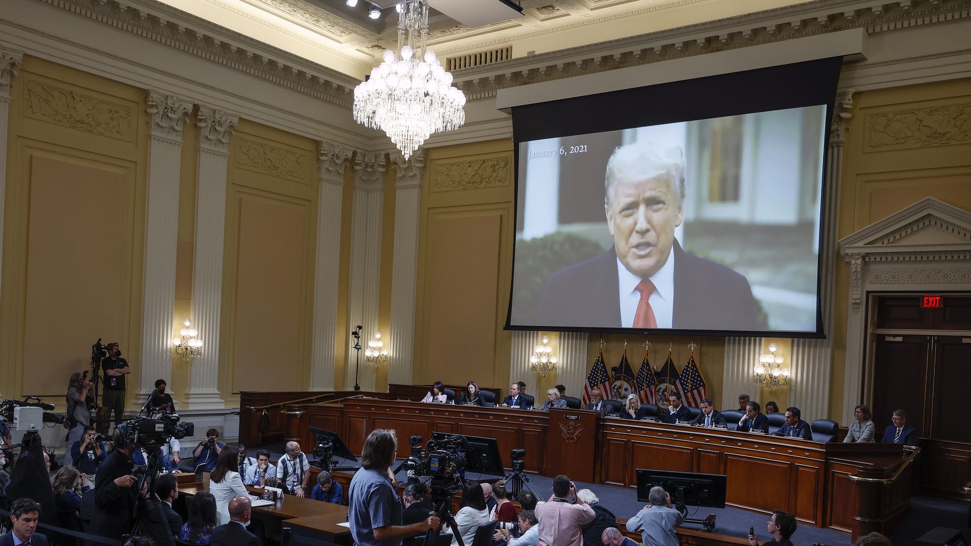 Photo of a projector showing Donald Trump on a screen that hangs over a row of Congress members sitting at the front of a hearing room