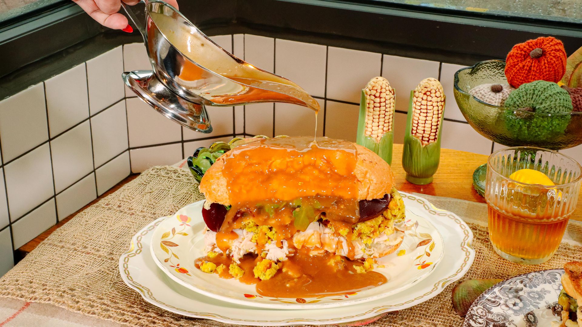 Hand with red nails pouring brown gravy from a silver gravy boat over a turkey sandwich with stuffing and cranberry sauce on a decorative plate, with corn-shaped salt and pepper shakers and knit pumpkins in the background.