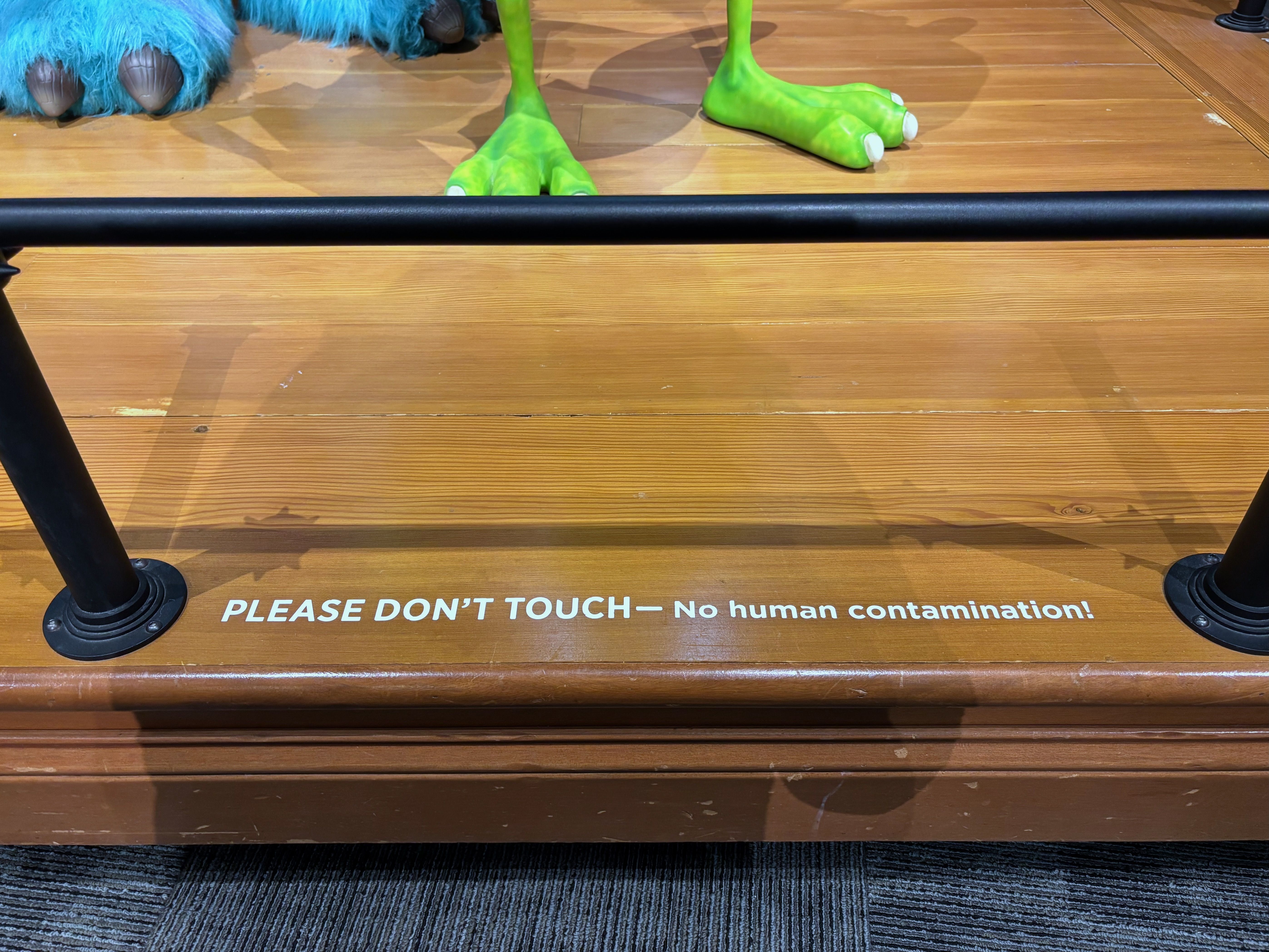 Sign on wooden floor with black railing saying PLEASE DON'T TOUCH — No human contamination, below feet of green and blue furry costume characters.