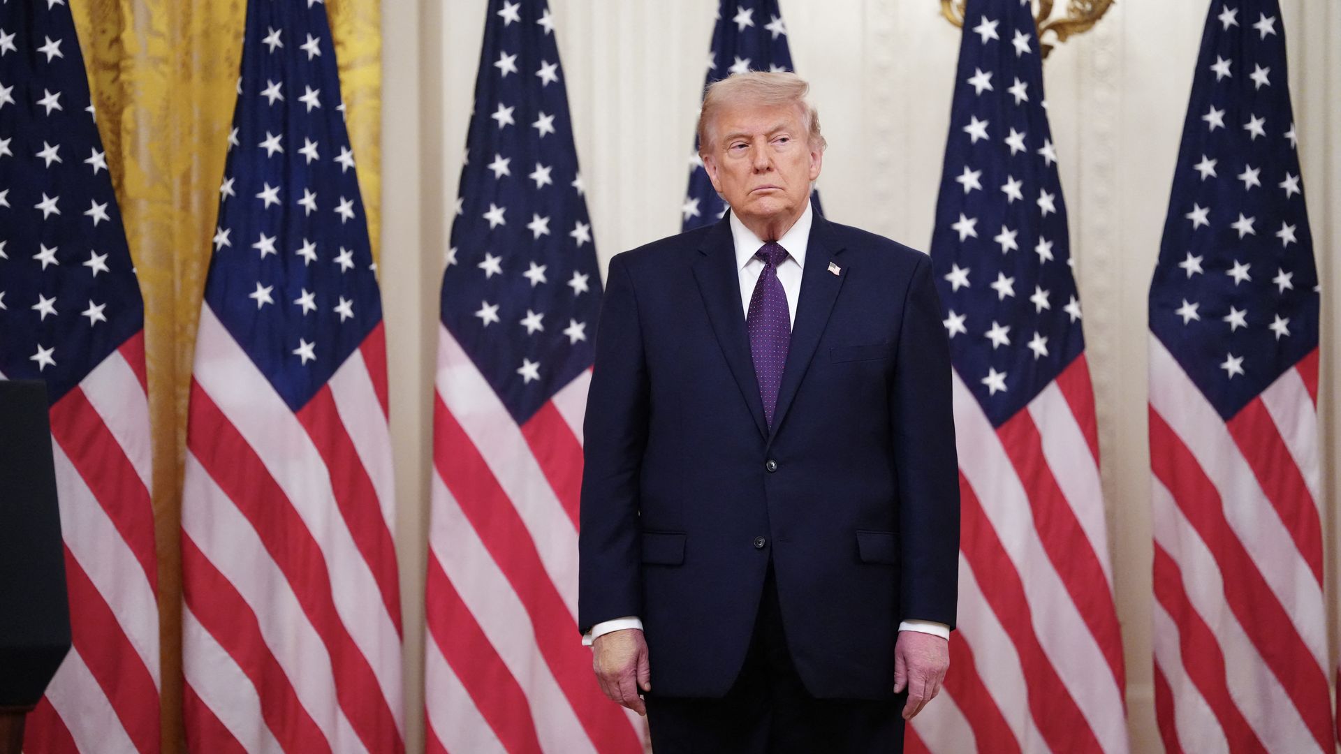 A man in a dark suit and purple tie stands before a row of American flags at a formal event, with a lapel pin and marble columns in the background.