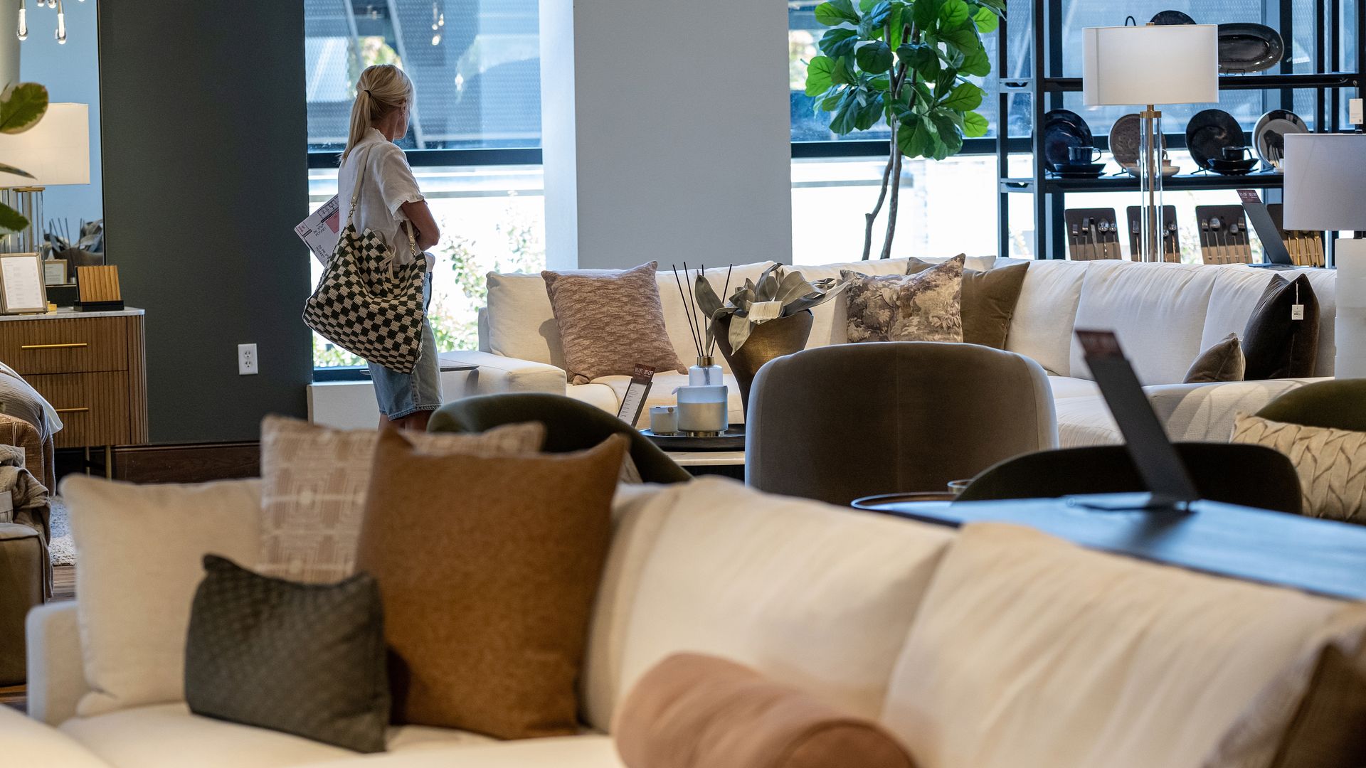 Woman with a checkered bag browsing a showroom filled with beige and brown couches, cushions, plants, lamps, and shelves with dishware and cutlery.
