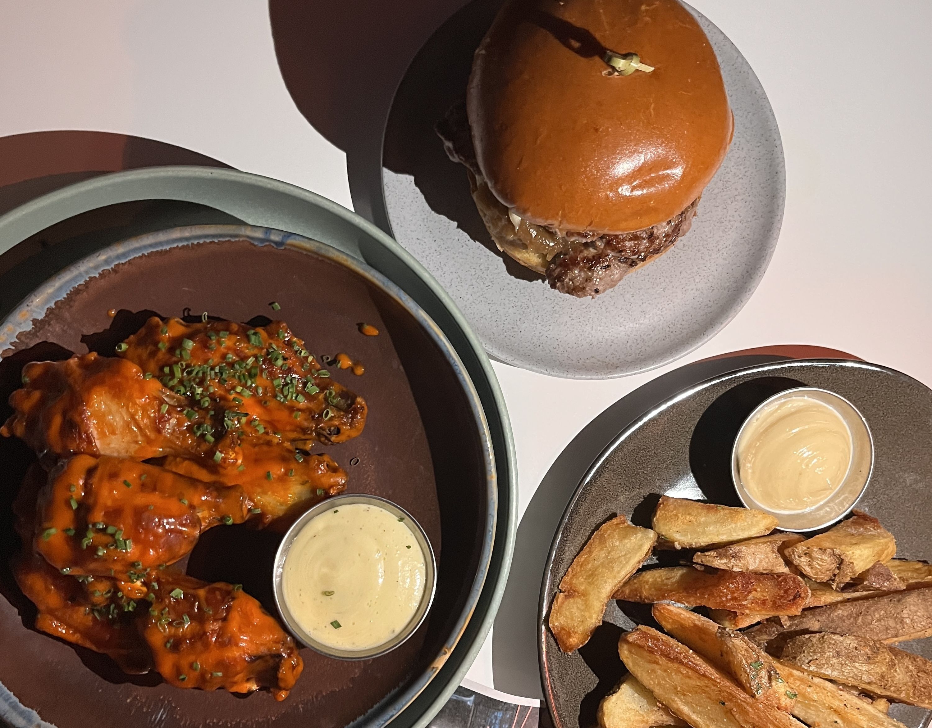Buffalo chicken wings with dipping sauce, a cheeseburger on a brioche bun, and thick-cut fries with dipping sauce on gray and brown plates on a white table.