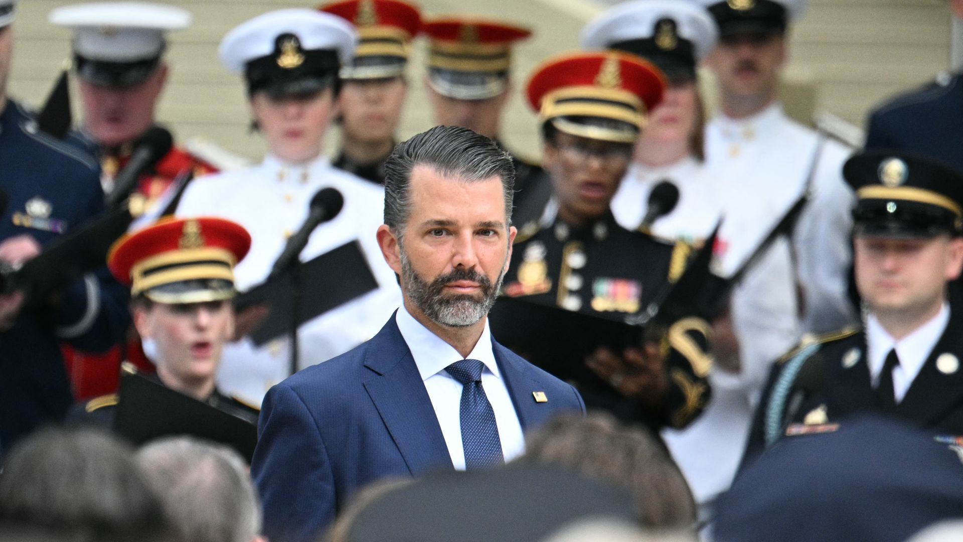 Centered man with a trimmed beard in a navy suit and blue tie at a formal ceremony; behind him, blurred uniformed military personnel in white, red, and dark dress uniforms with peaked caps.