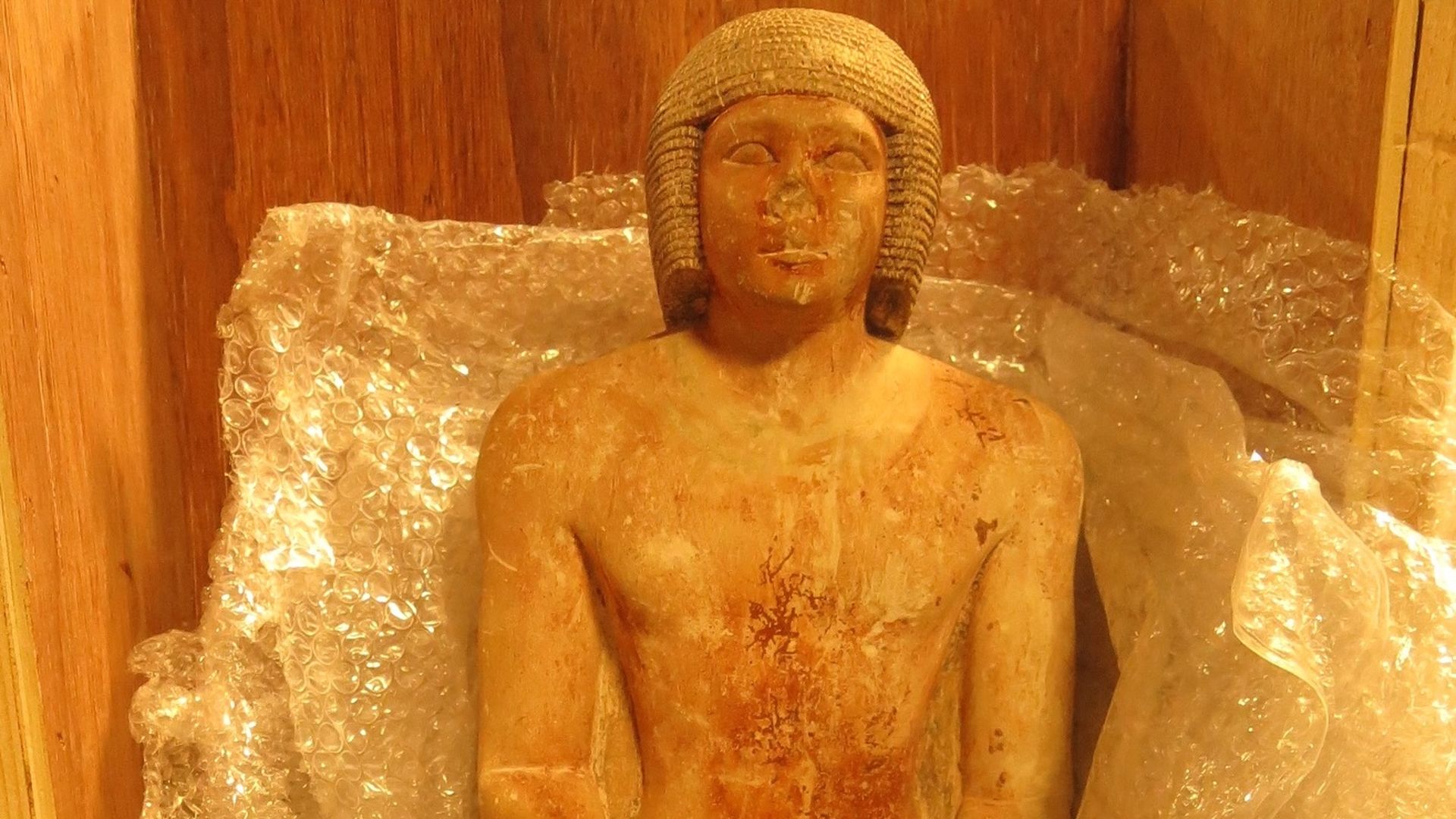 The Old Kingdom Limestone Funerary Statue, a browning ancient Egyptian artifact depicting an ancient Egyptian person, with the nose fallen off, lying in bubble wrap in a wooden case.