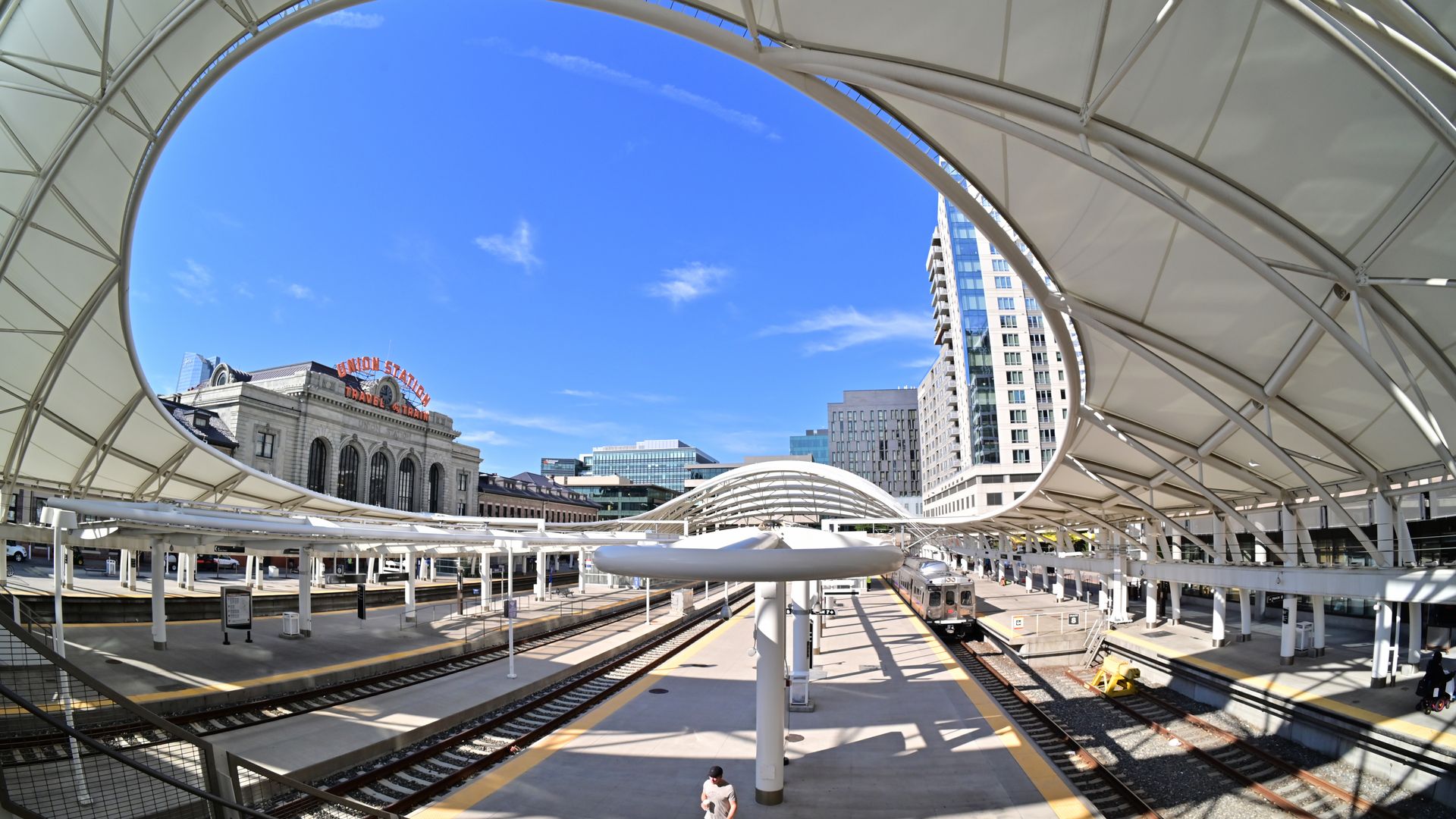 A large train platform with a white canopy, and several train tracks with a yellow lining. In the middle,  large structure with the words UNION STATION sits in the middle, while apartment buildings stand high across it. 