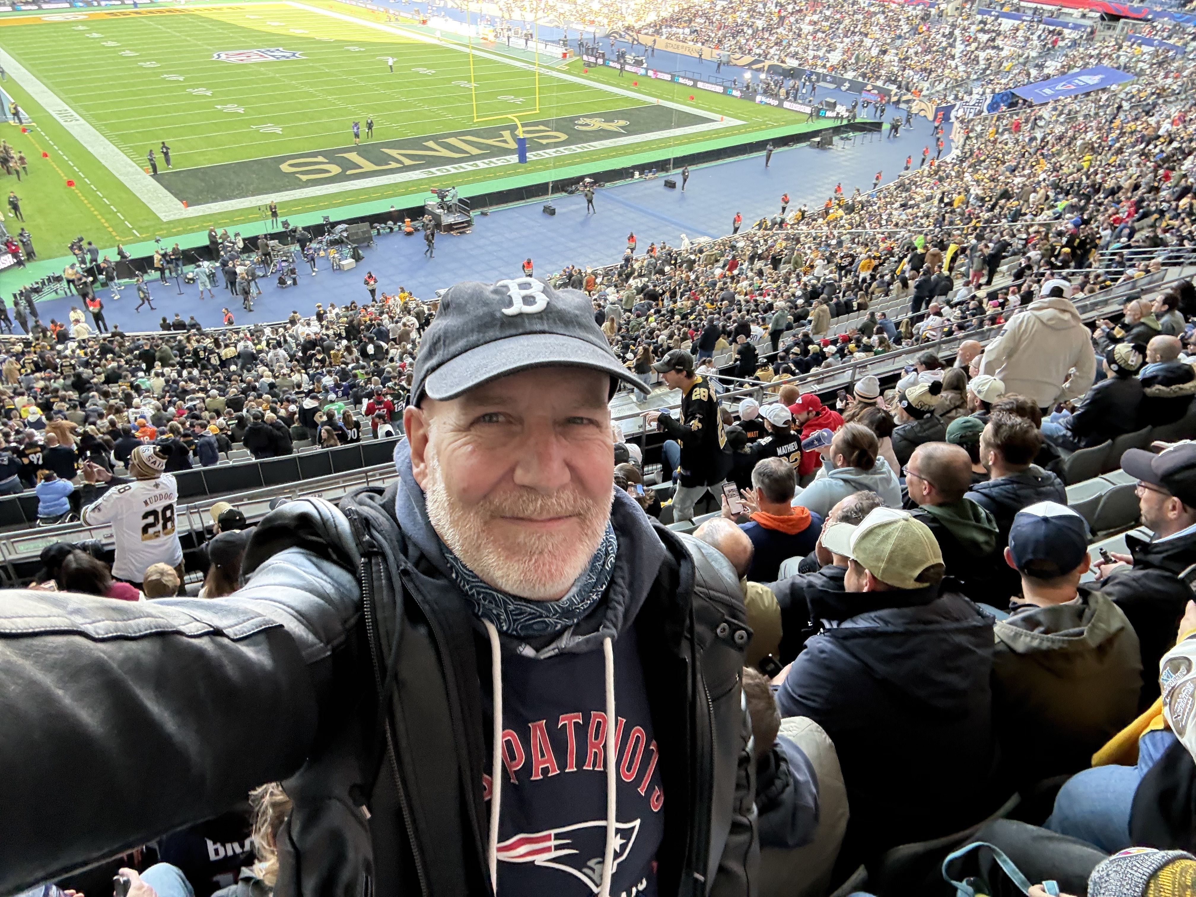 Selfie of a smiling man wearing a gray Patriots cap and jacket, in the stands at a packed football stadium. The field shows the Saints end zone, yellow goalposts, and cheering crowd.