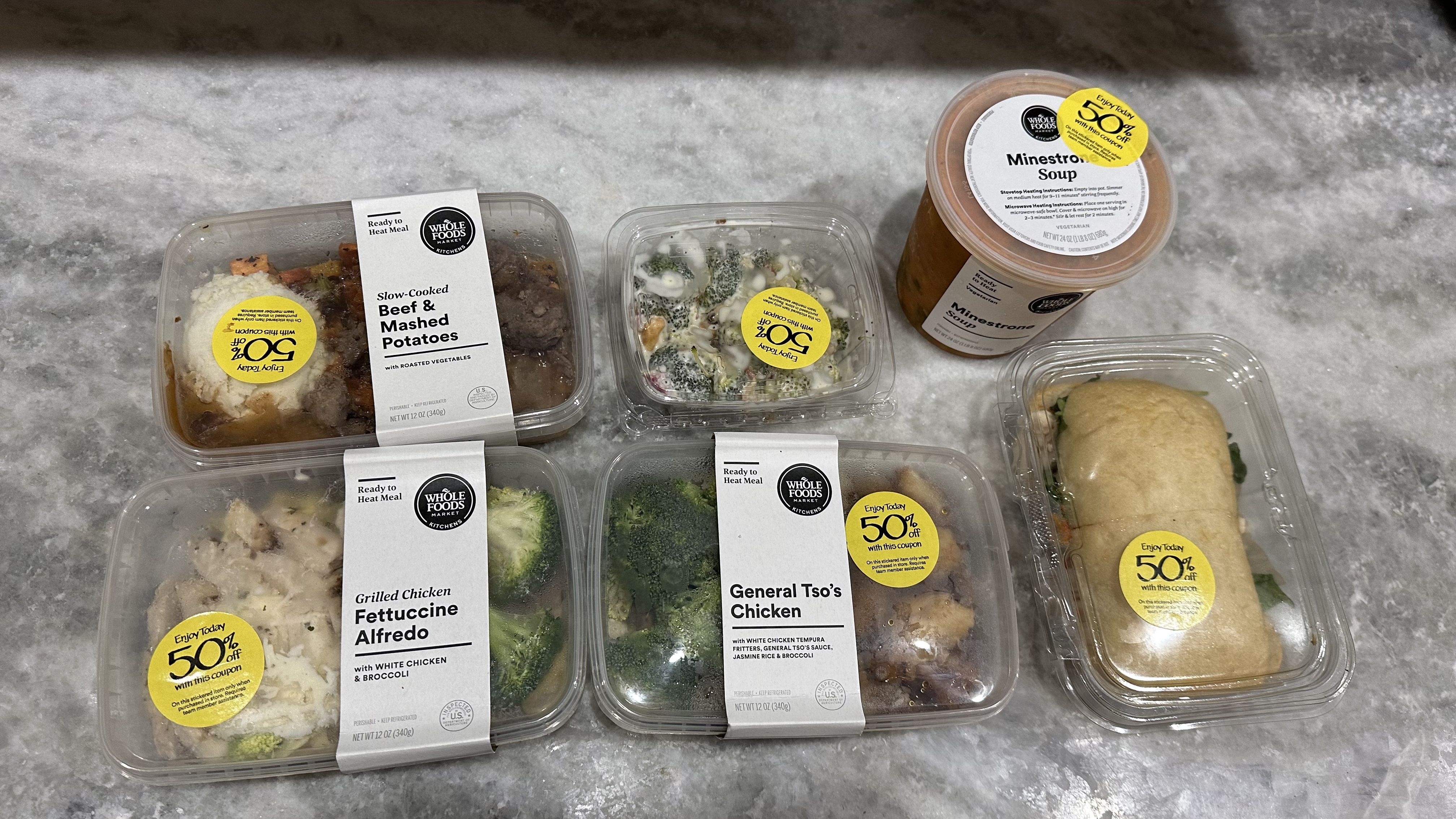 Prepared foods from Whole Foods on a marble counter. The foods are beef and mashed potatoes, fettucine alfredo, General Tso's Chicken, a sandwich and a quart of soup.
