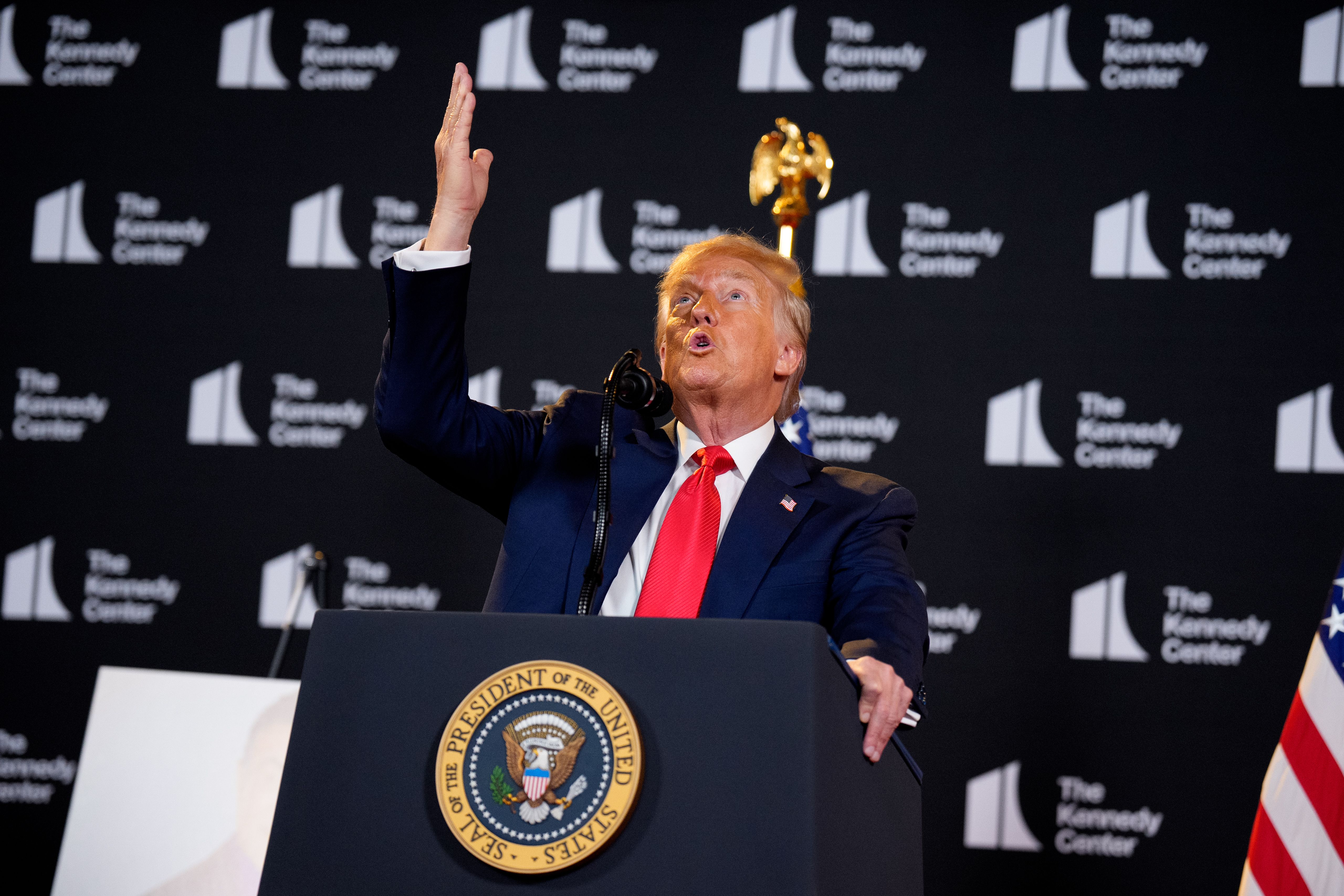 A man in a navy suit with a red tie speaking at a podium with the United States presidential seal, raising one hand, with a black backdrop repeating "The Kennedy Center" logo.