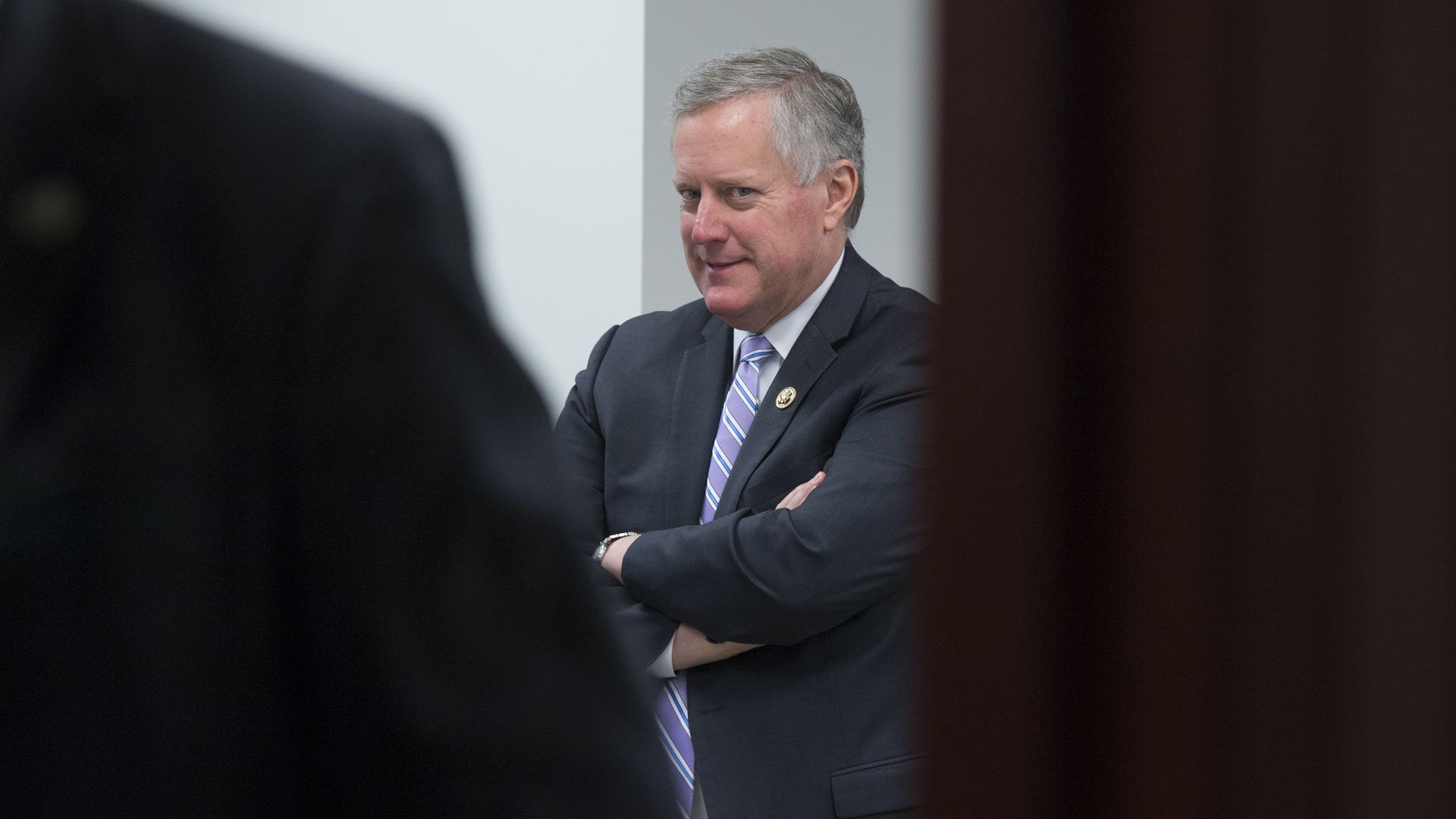 Rep. Mark Meadows stand in a dark suit with his arms crossed with two dark figures on either side in the foreground