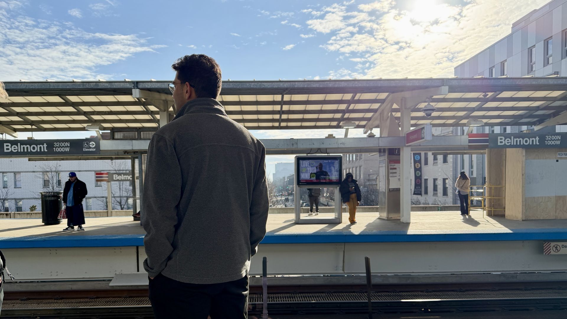 A man in a gray jacket stands with his back to the camera on a sunlit Belmont station platform. Blue sky overhead, sheltered walkway, and other commuters visible in the background.