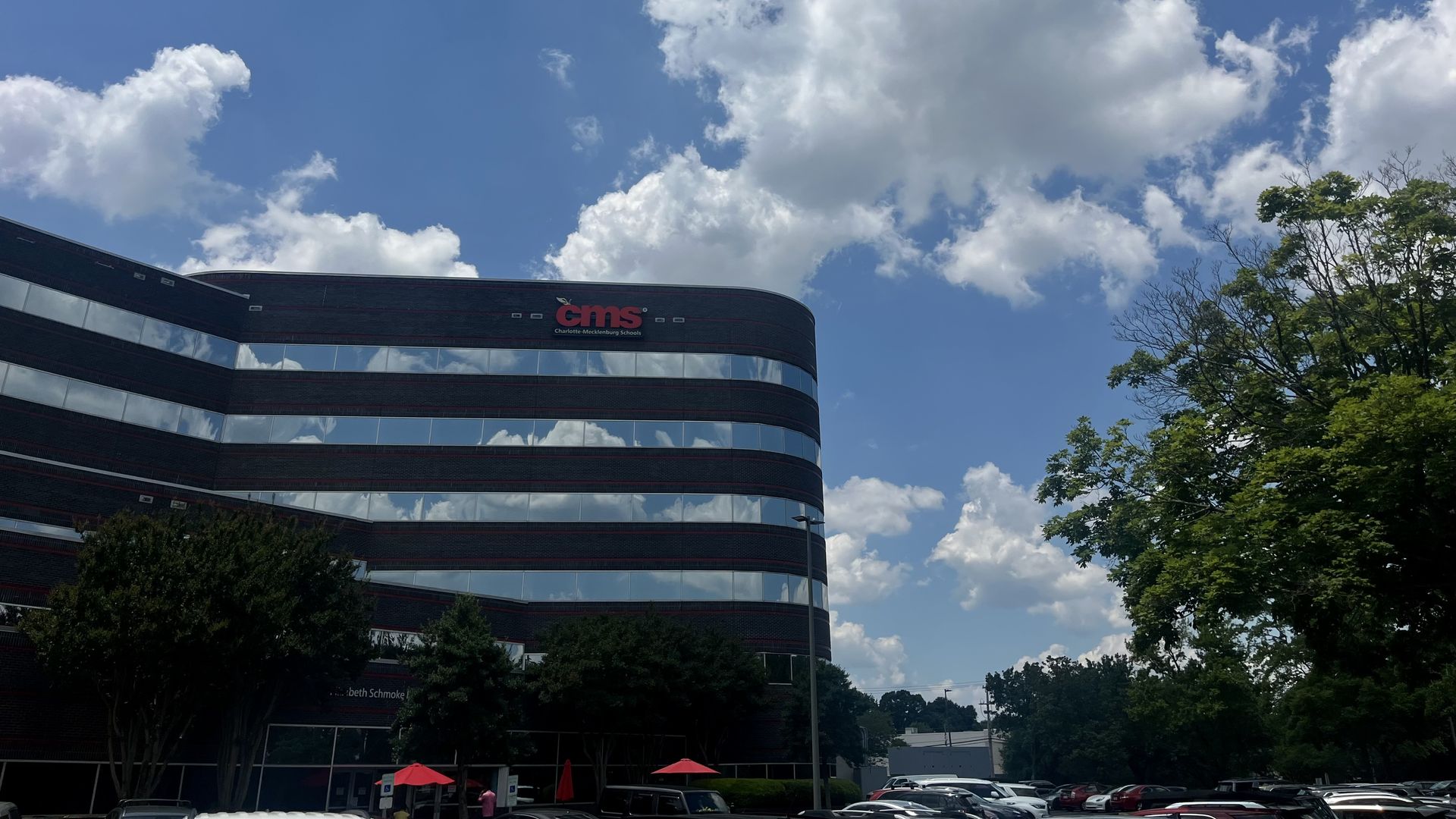Office building with "cms" and "Charlotte-Mecklenburg Schools" logo on a sunny day with blue sky and white clouds, trees around, and a parking lot with cars and red umbrellas.