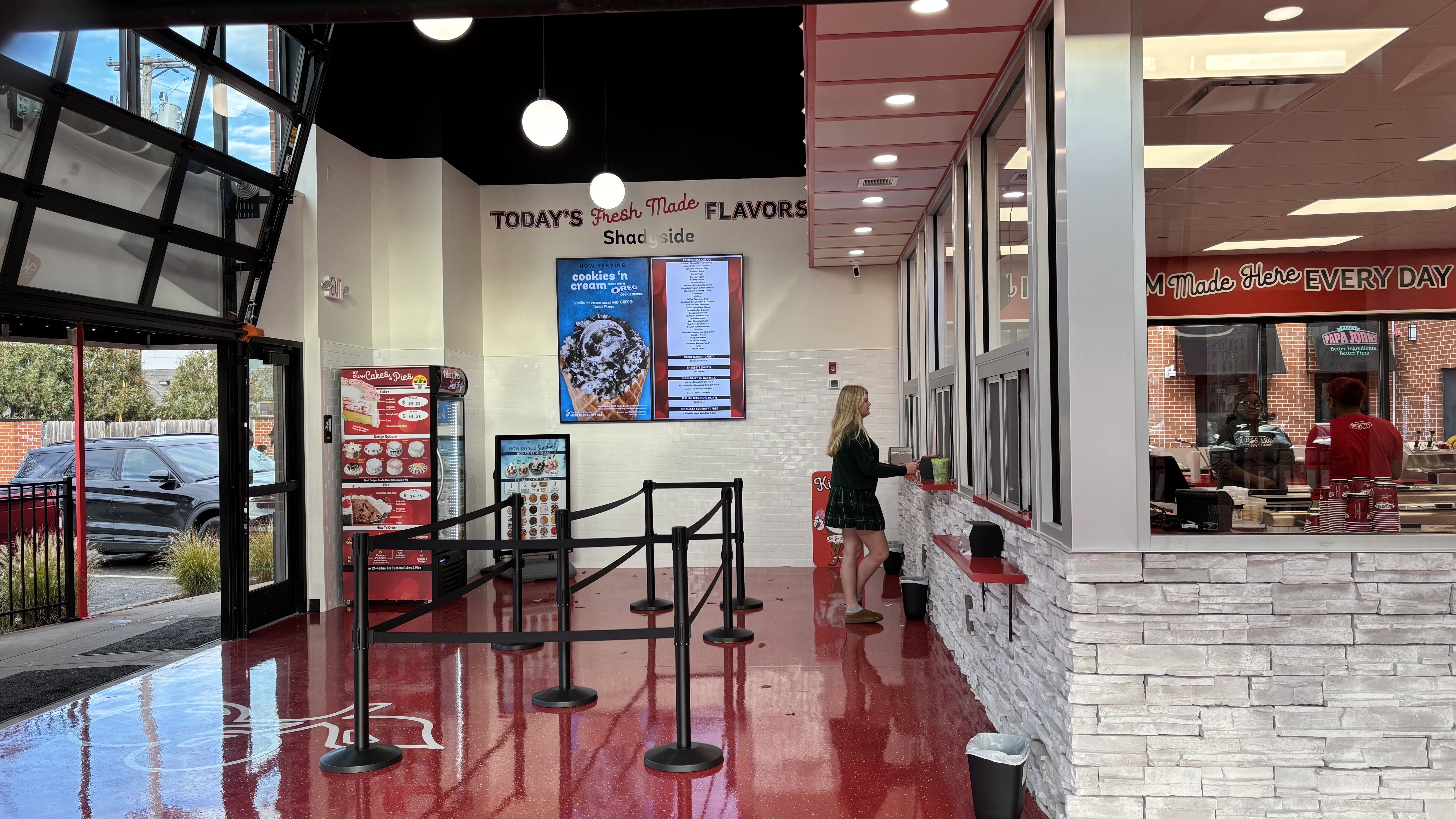 Interior of an ice cream shop with shiny red floor, black queue stanchions, white stone counter, and a woman ordering at the window. Menus and signs advertise flavors and cakes.