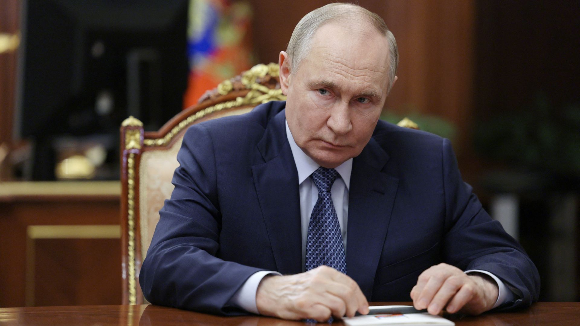 A man in a dark blue suit and patterned blue tie sits at a polished wooden table, looking serious, in an ornately decorated room with a blurred computer and colorful flag in the background.