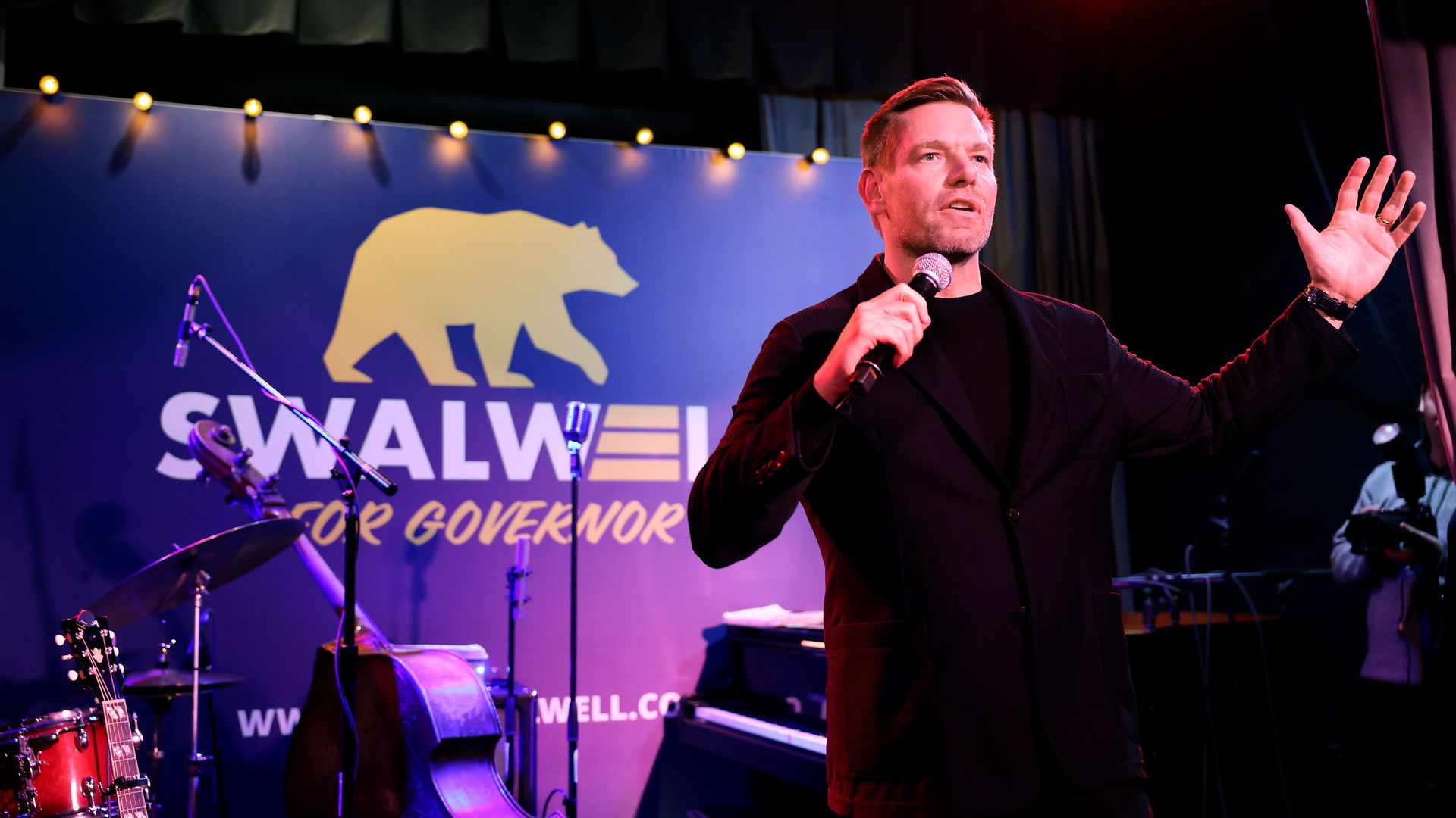 A man in a black blazer speaks into a microphone on a stage with a blue backdrop showing a yellow bear and Swalwell for Governor; drums and a guitar sit left, under purple and red stage lights.