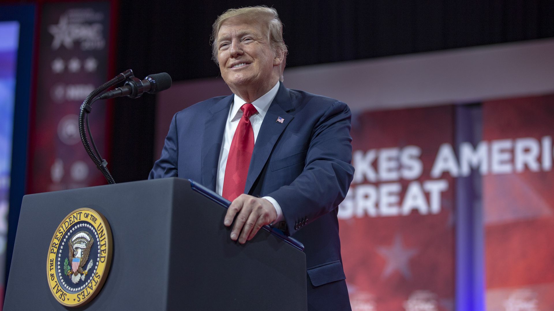 In this image, Trump stands and smiles behind a podium at CPAC.