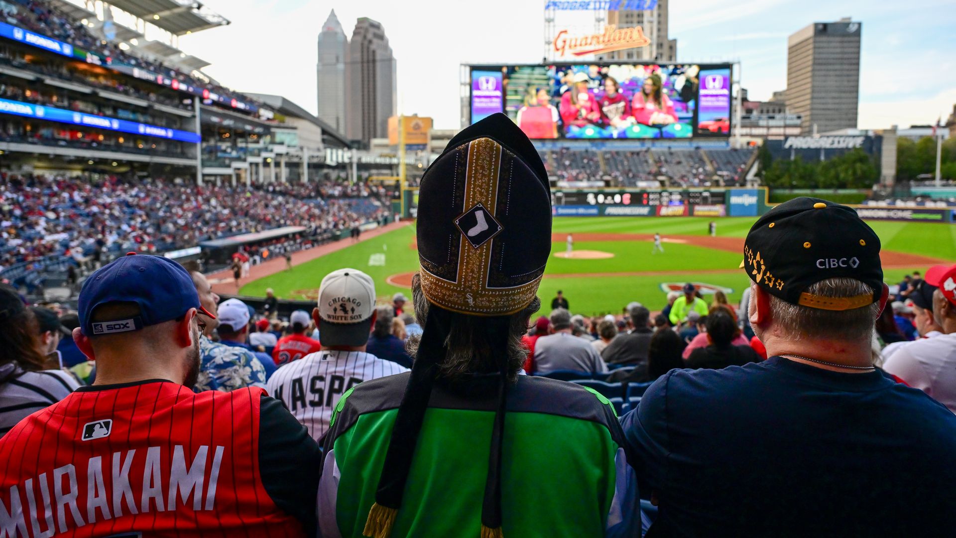View from behind a crowded baseball stadium: a fan in a tall black and gold feather headdress dominates the foreground, while the big video board and city buildings loom in the background.