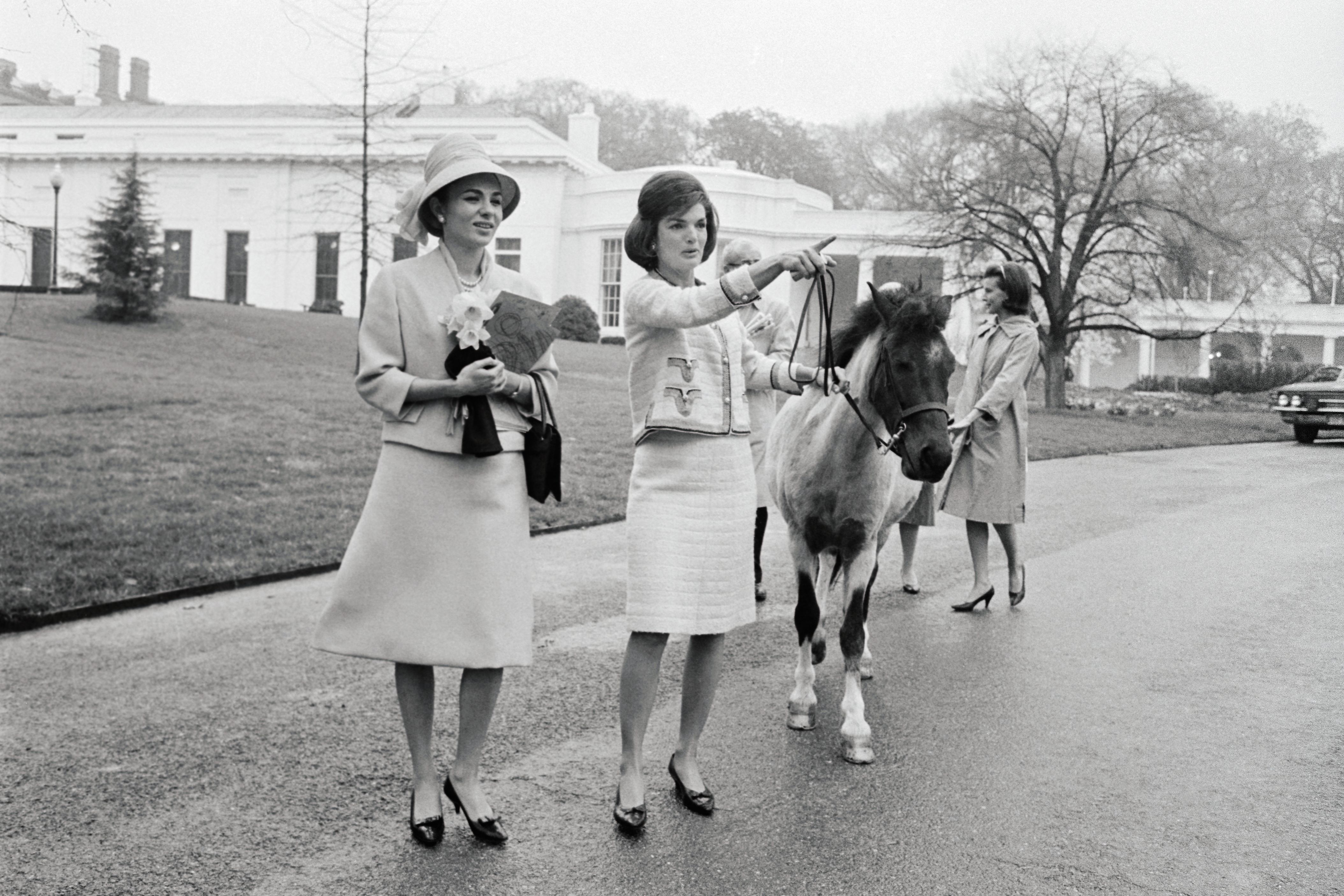 A Empress Farah, wearing a hate and lightc-colored skirt suit, clutches her hand as first lady Jacqueline Kennedy, with dark, shoulder-length hair and wearing a light colored suit, points and carries the reins of a horse she's leading in the White House grounds, near the East Colonnade in 1962.