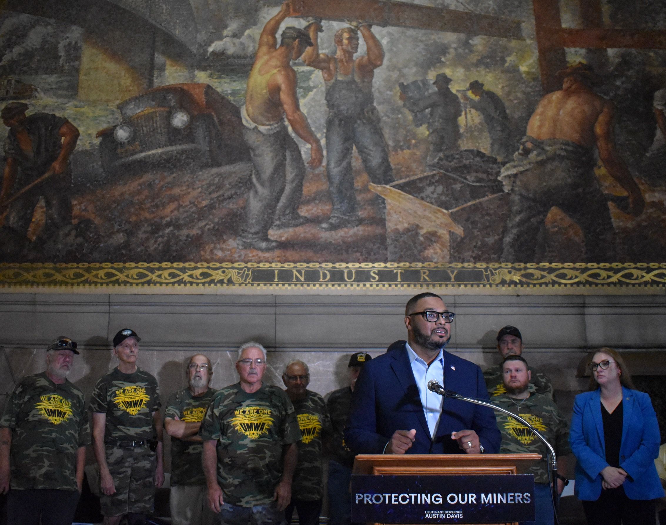 A man in a blue suit speaks at a podium reading "Protecting Our Miners" with a group of people in camo shirts behind him, standing under a mural depicting industrial workers.