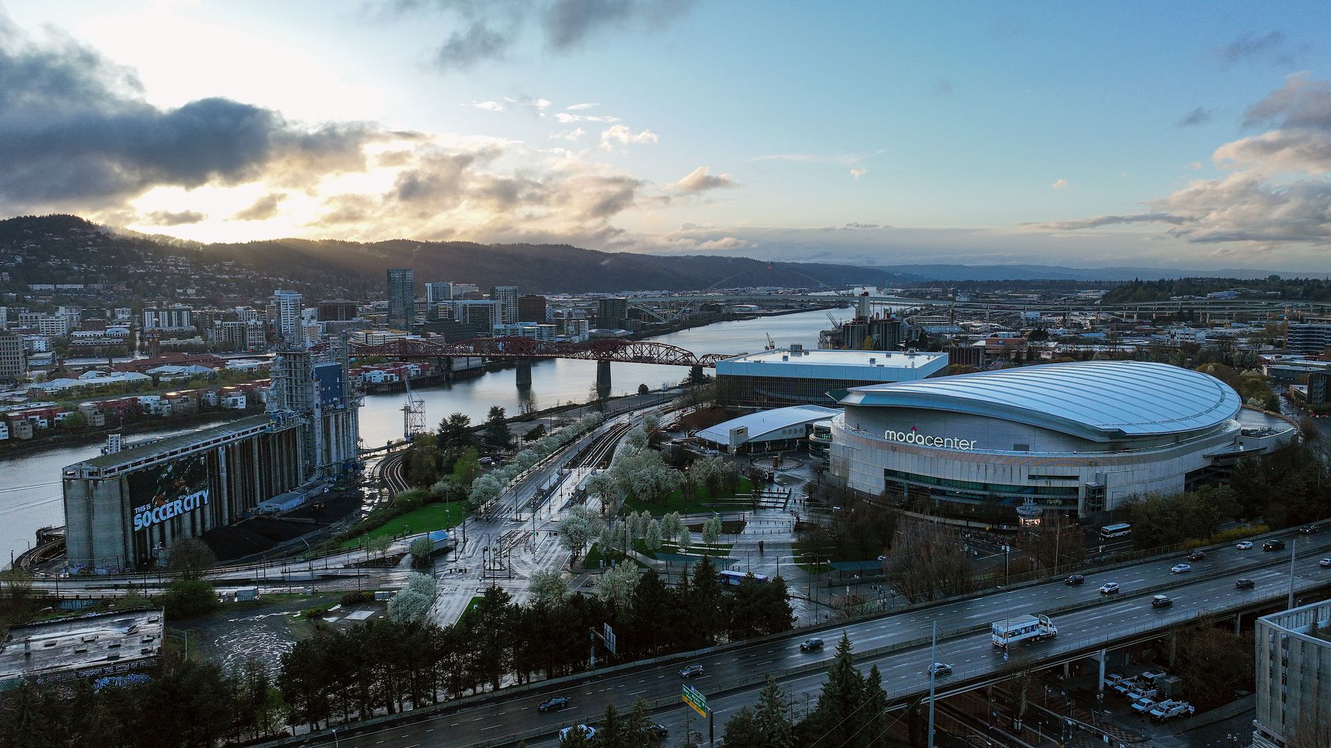 Wide cityscape with a winding river, multiple bridges, and a large curved arena labeled "Moda Center" in the foreground. Downtown buildings, hills, and a sunset sky.