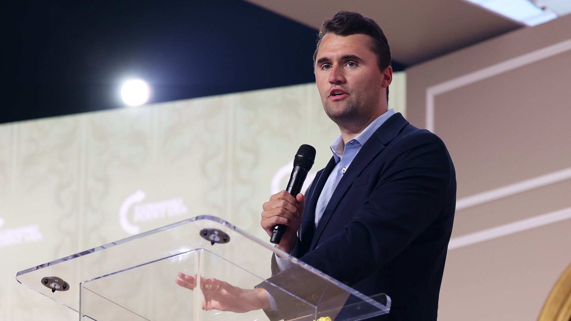 Charlie Kirk, wearing a blue suit and holding a microphone in one hand while placing his other on a translucent podium, speaks in front of a white and gray wall.