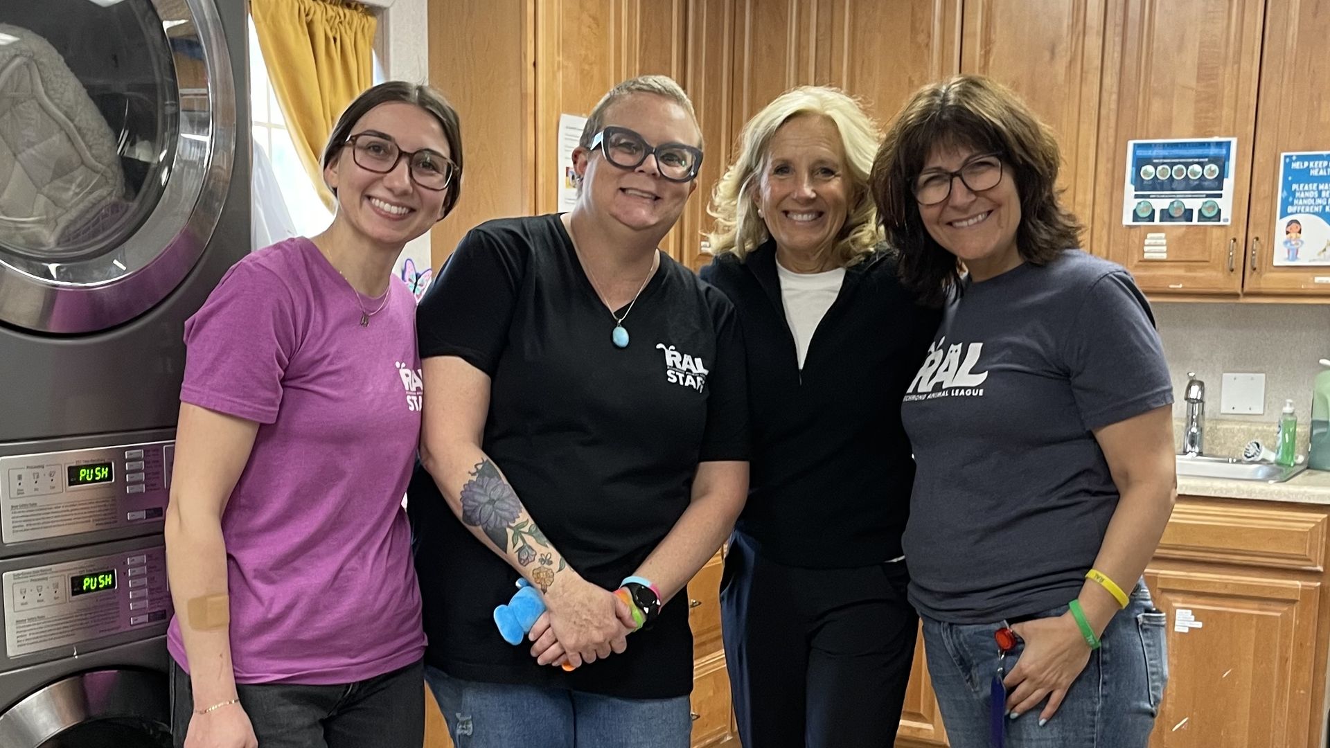 Four women stand together in a laundry room, smiling at the camera. A large front-loading washer sits to the left; wooden cabinets and posters line the back wall. They wear casual shirts and sneakers.
