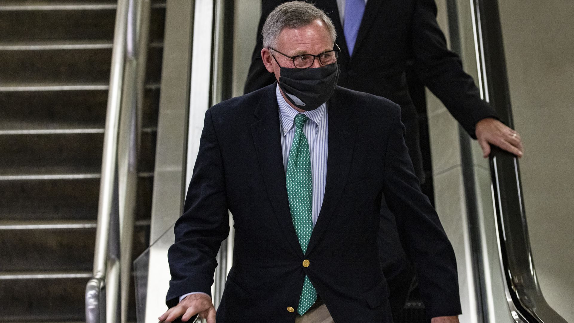 Richard Burr uses an escalator while wearing a face mask and a suit and tie
