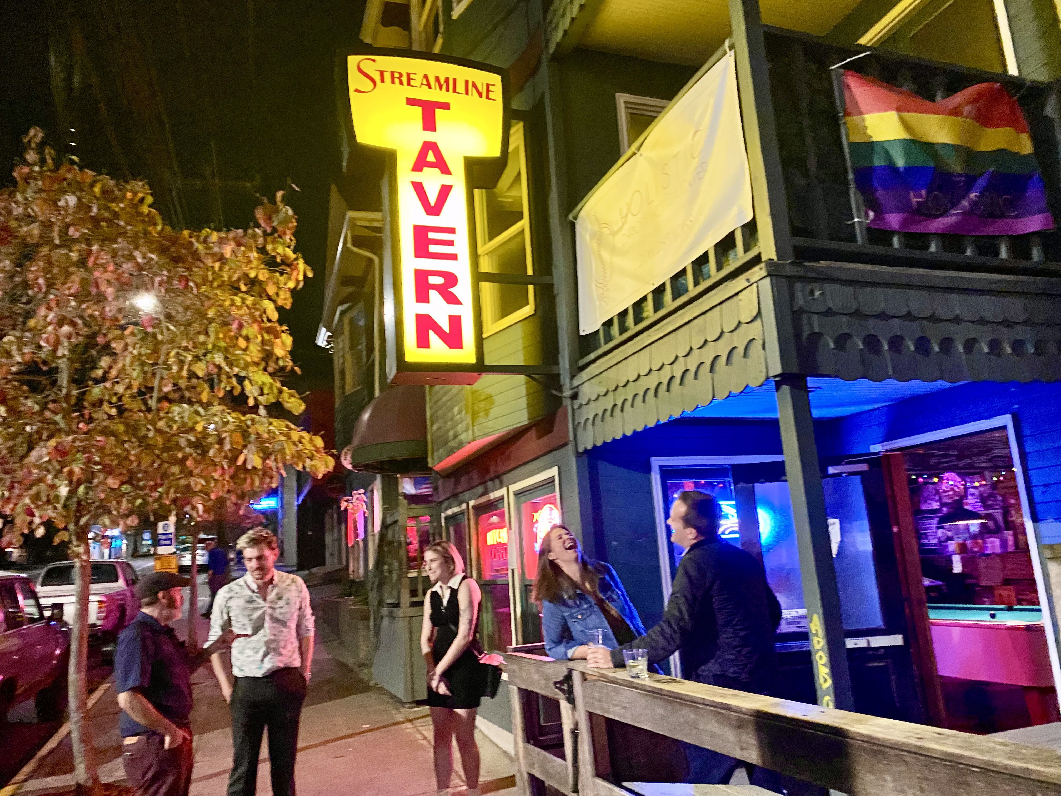 People stand on a sidewalk and on an outdoor patio at a bar, talking, with a yellow sign that says Streamline Tavern above. 