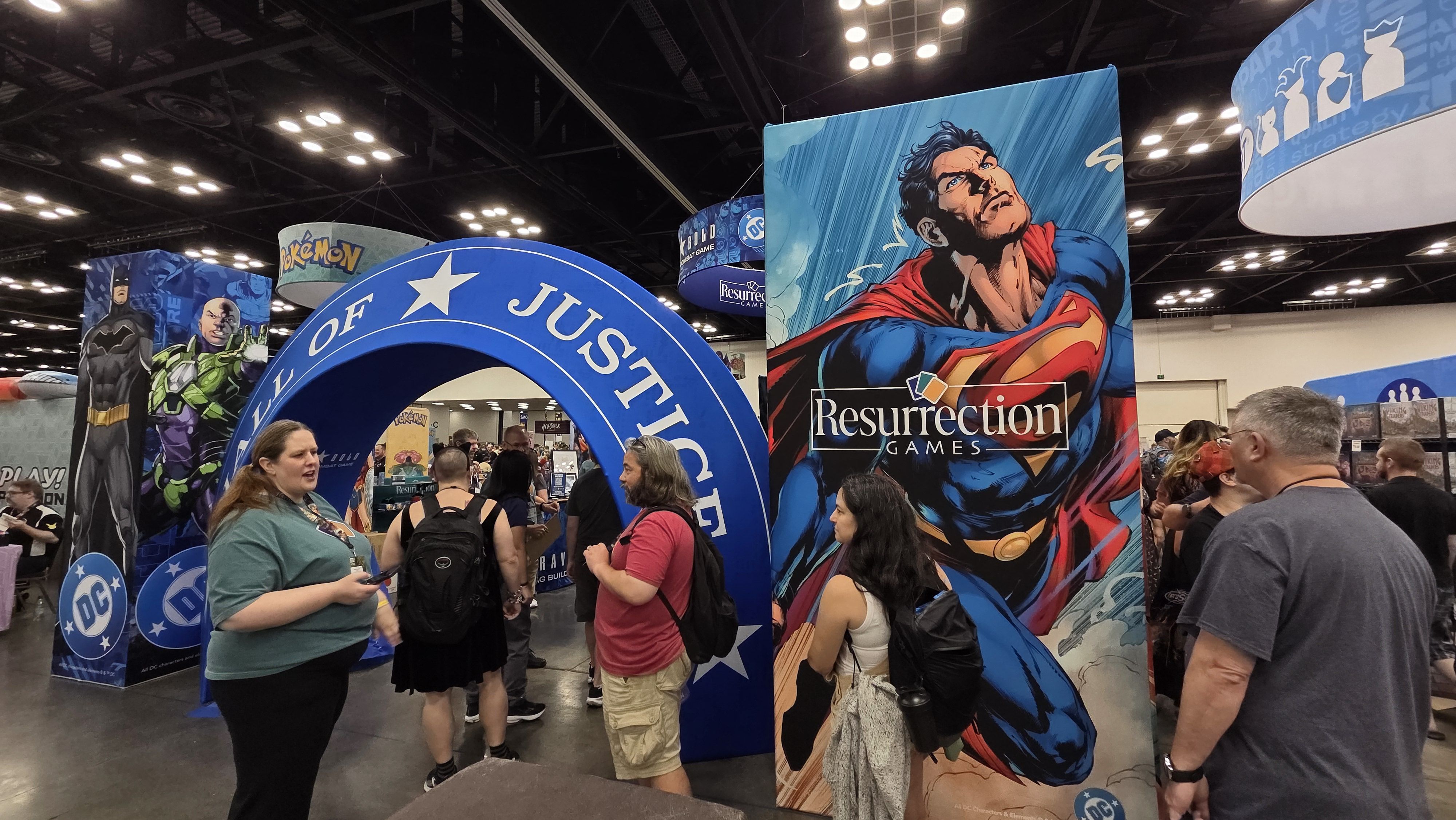 Busy convention hall with large DC Comics displays: Batman and Green Lantern cutouts on the left, a giant Superman panel with "Resurrection Games" to the right, and visitors browsing booths.