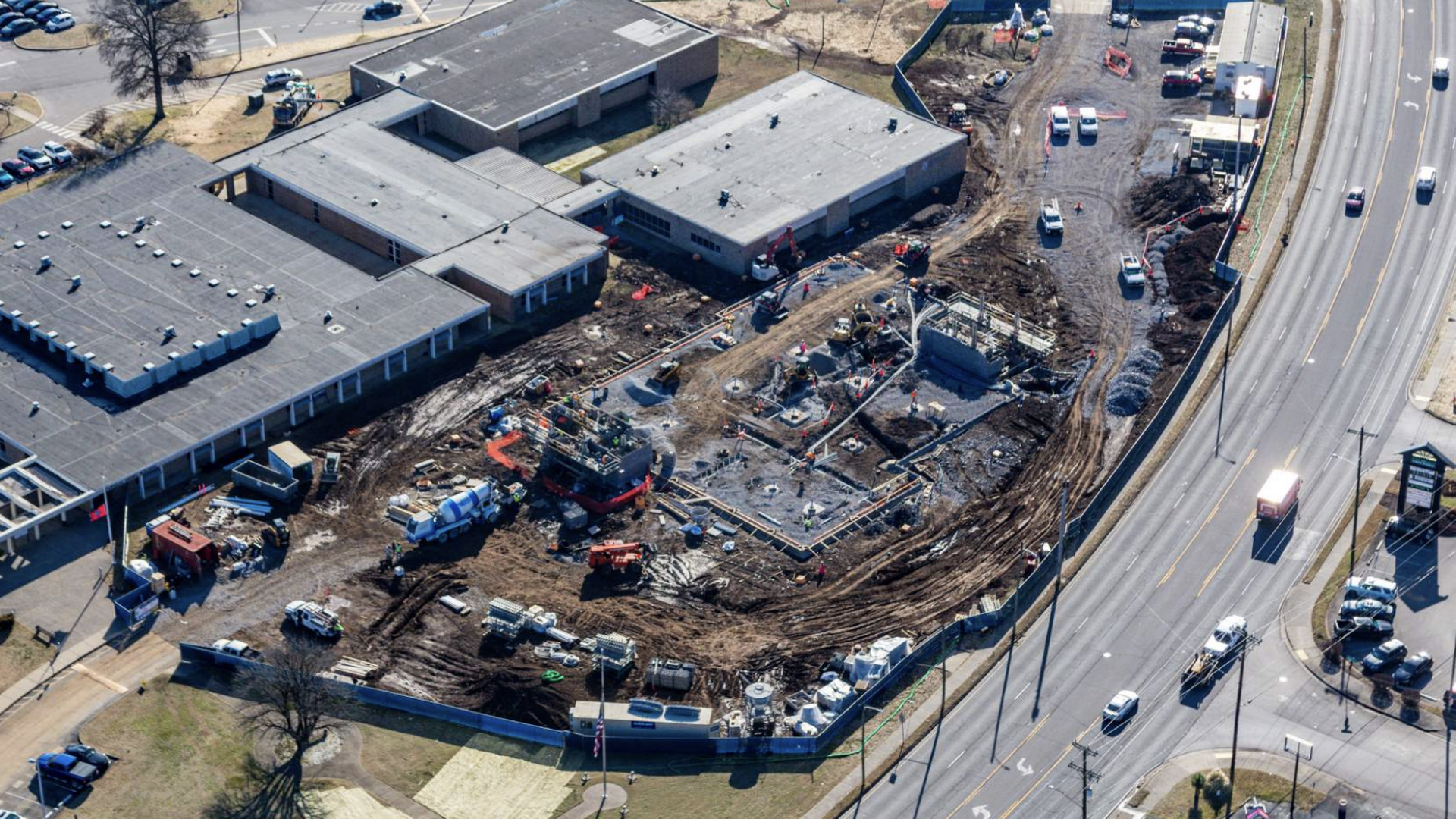 Aerial view of a construction site next to large gray buildings with flat roofs, surrounded by roads and parked cars, with construction vehicles and workers active on muddy ground.