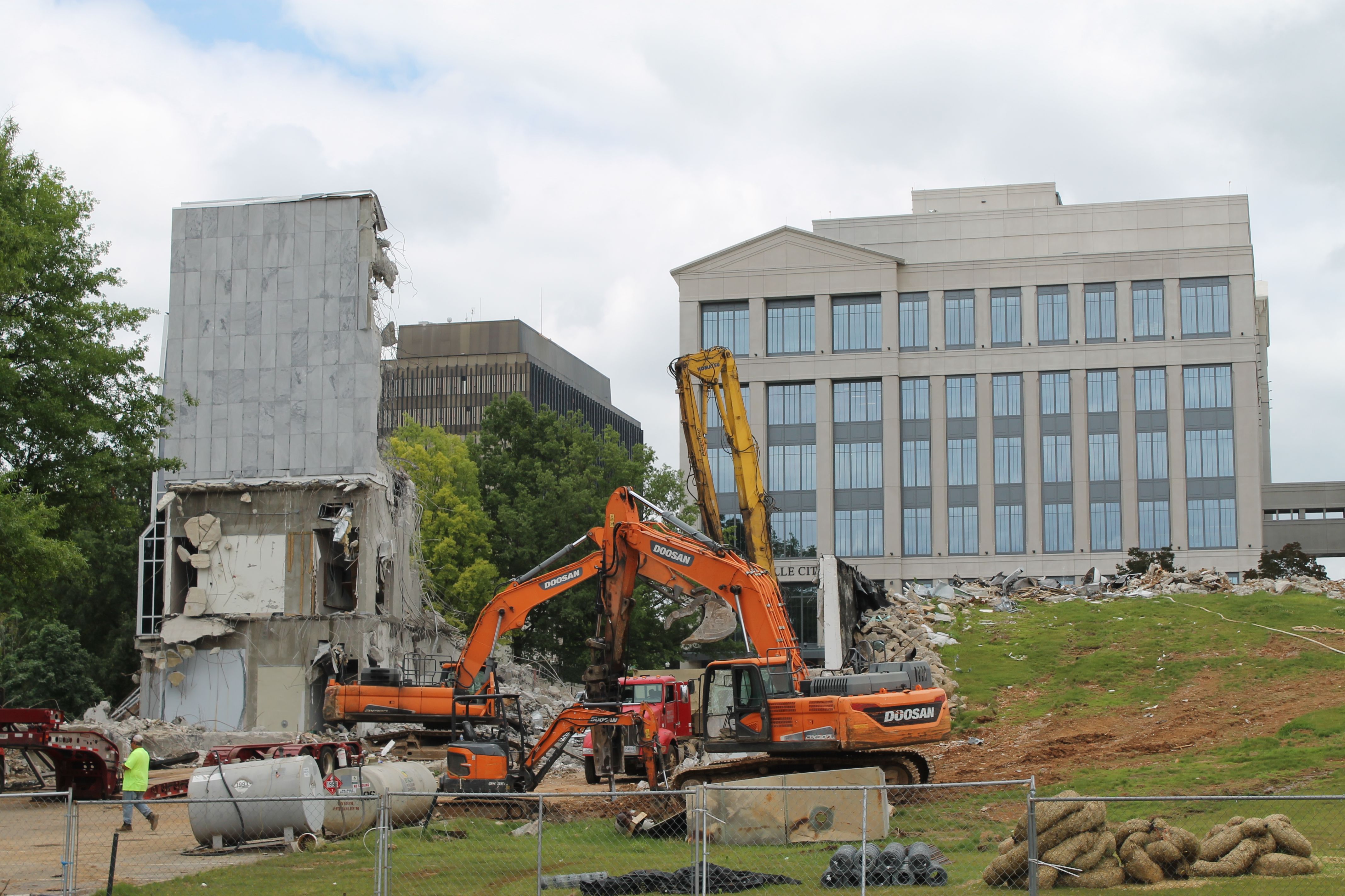 Construction site with partially demolished building, orange and yellow Doosan excavators, a worker in a neon yellow shirt, grassy area, and modern multi-story building in the background under a cloudy sky.