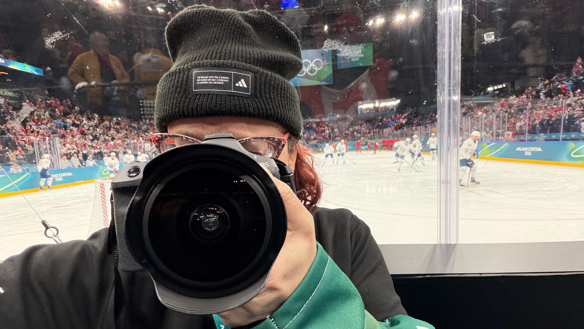 Photographer in black beanie with Adidas logo aiming camera at ice hockey game; players in white and red jerseys play on ice rink with crowd and Olympic rings banner in background.