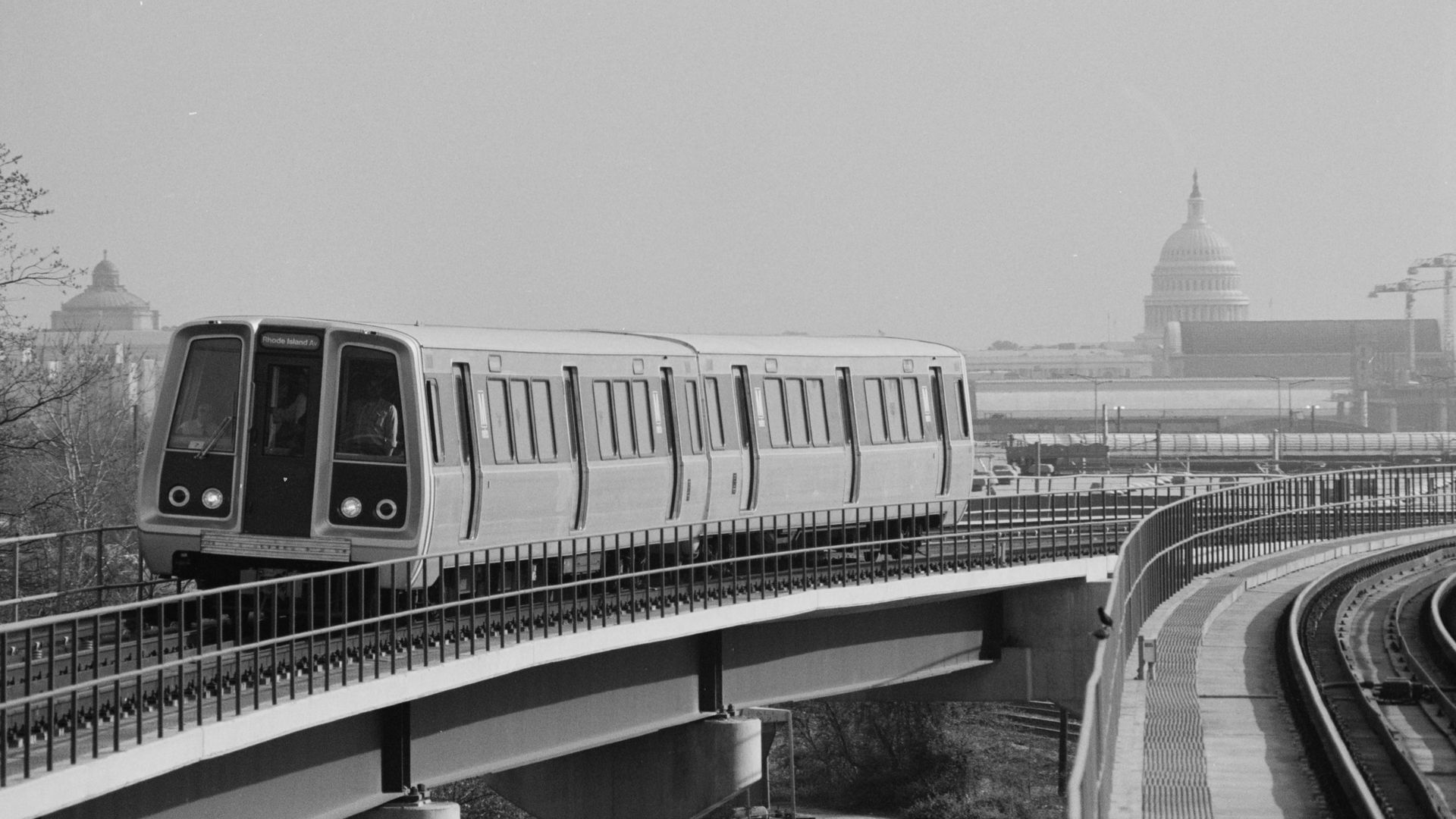 Monochrome image of a modern metro train on an elevated curved track with railing, moving left to right, as the U.S. Capitol dome and city skyline loom in the hazy background.