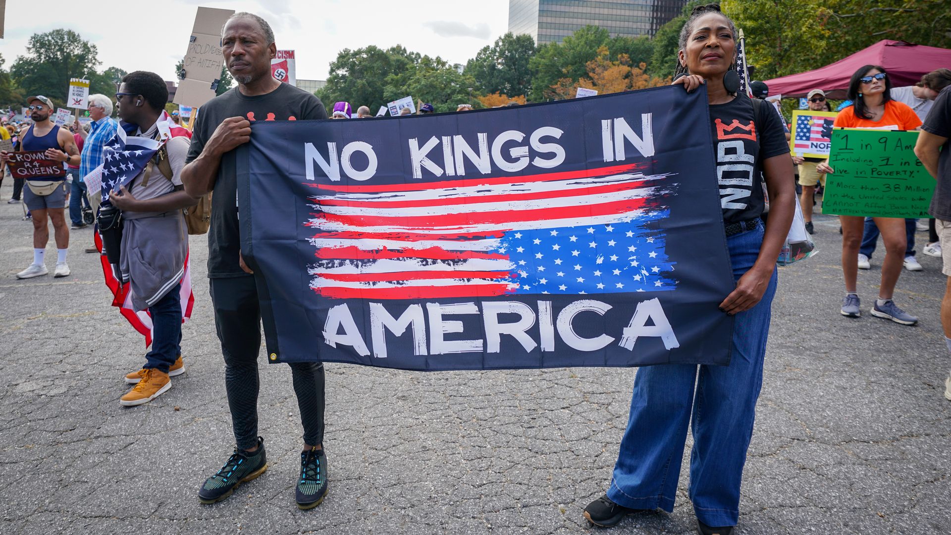 Two people hold a flag that reads "NO KINGS IN AMERICA," with the American flag drawn upside down.