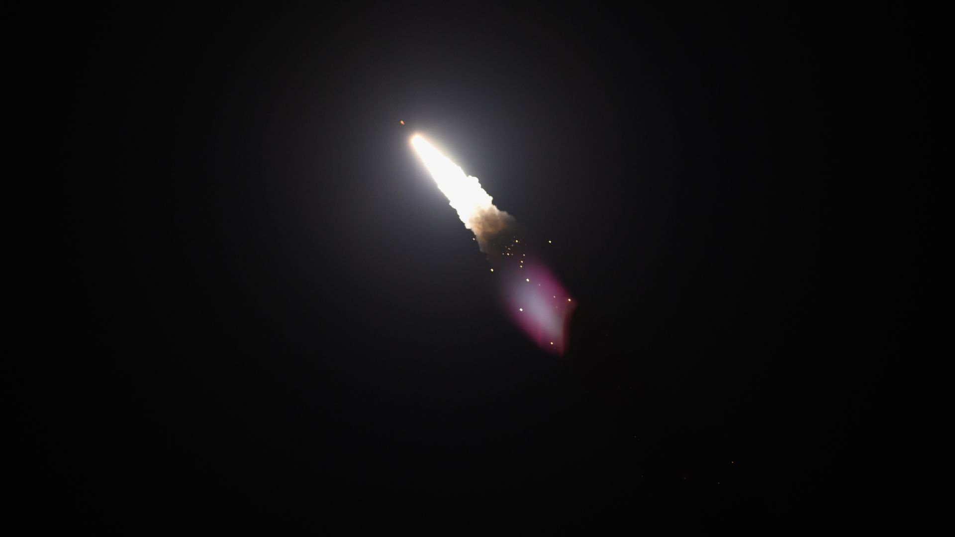A large missile soars through the night sky. Its trailed by bright smoke and a purple glare.