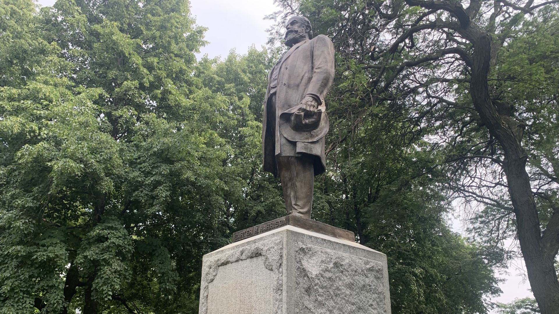 Bronze statue of a man holding a hat on a tall stone pedestal with a plaque in a park surrounded by green trees and parked cars in the background.