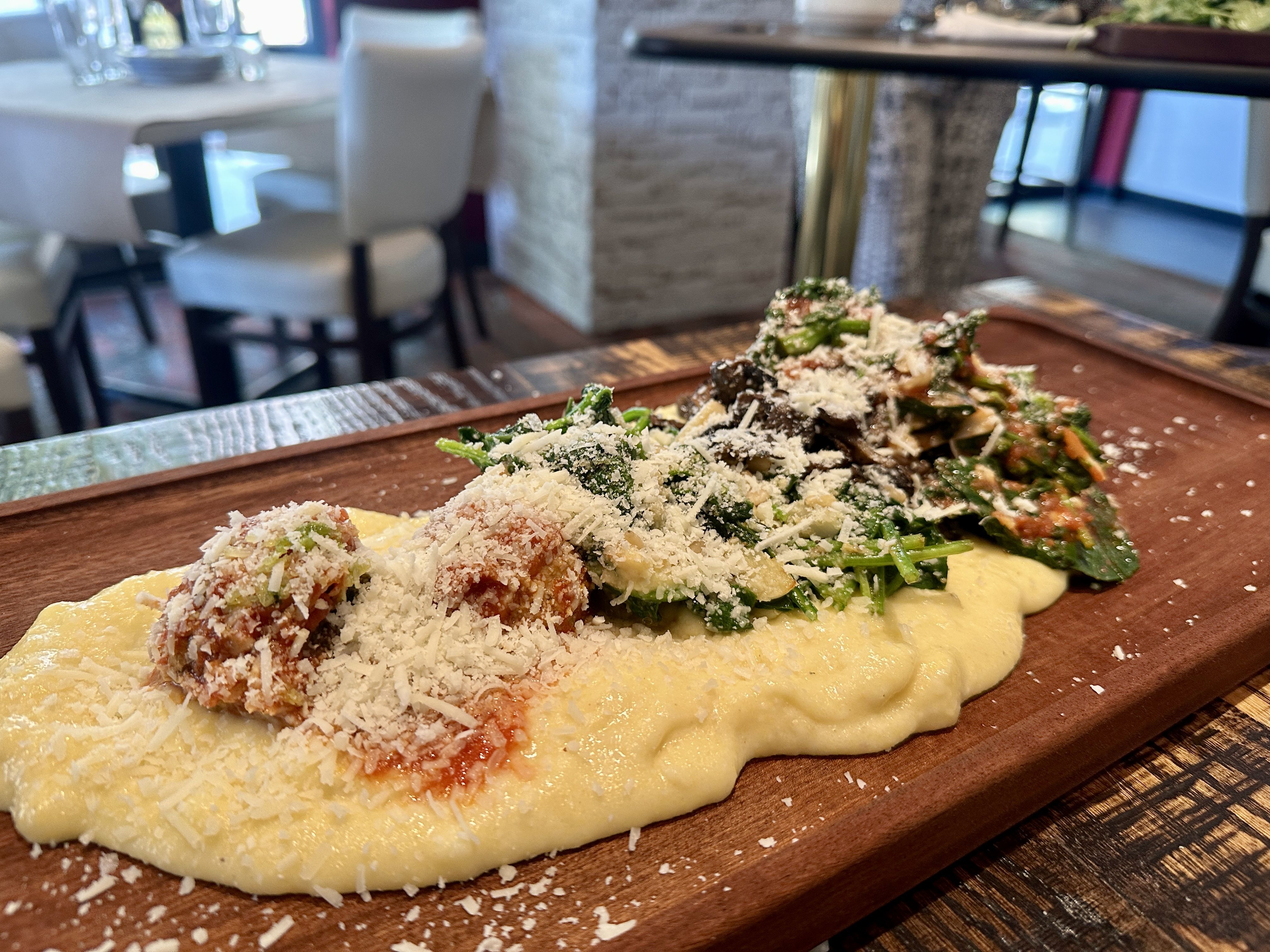 Close-up of a wooden platter with creamy yellow polenta topped with sautéed spinach, mushrooms, tomato sauce, and grated cheese in a restaurant setting with white chairs and tables blurred in background.