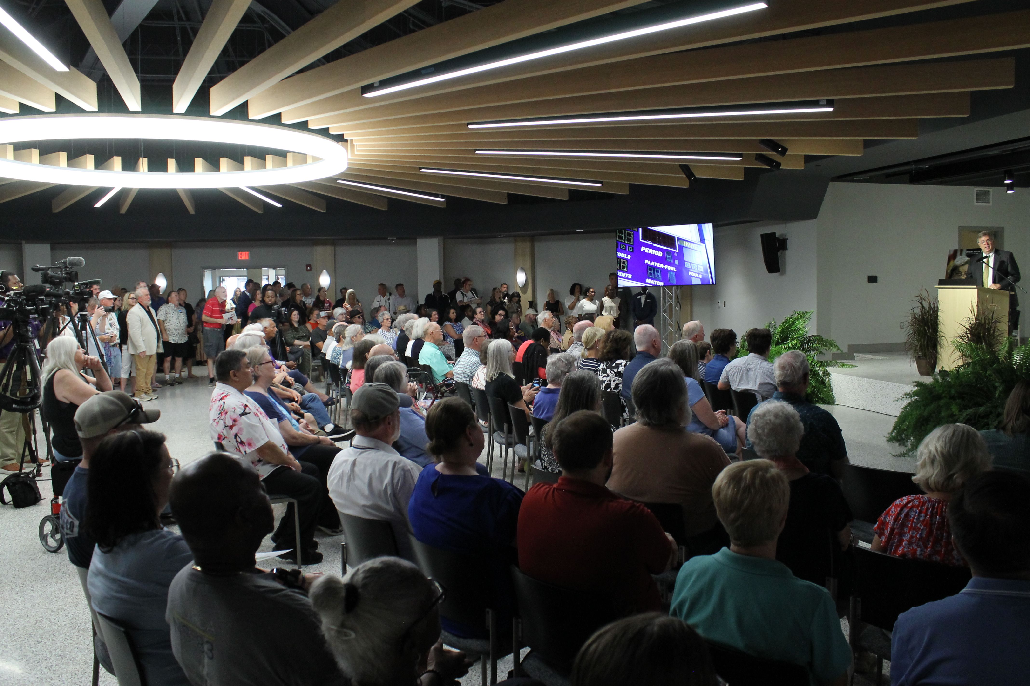 Audience seated in a modern event hall watches a speaker at a podium on the right; a large circular bright chandelier and wooden beam ceiling span overhead, with cameras along the left.