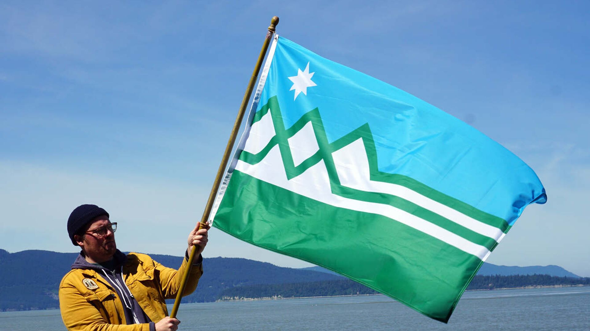 A man holds a blue, green and white flag with mountains and a star. 