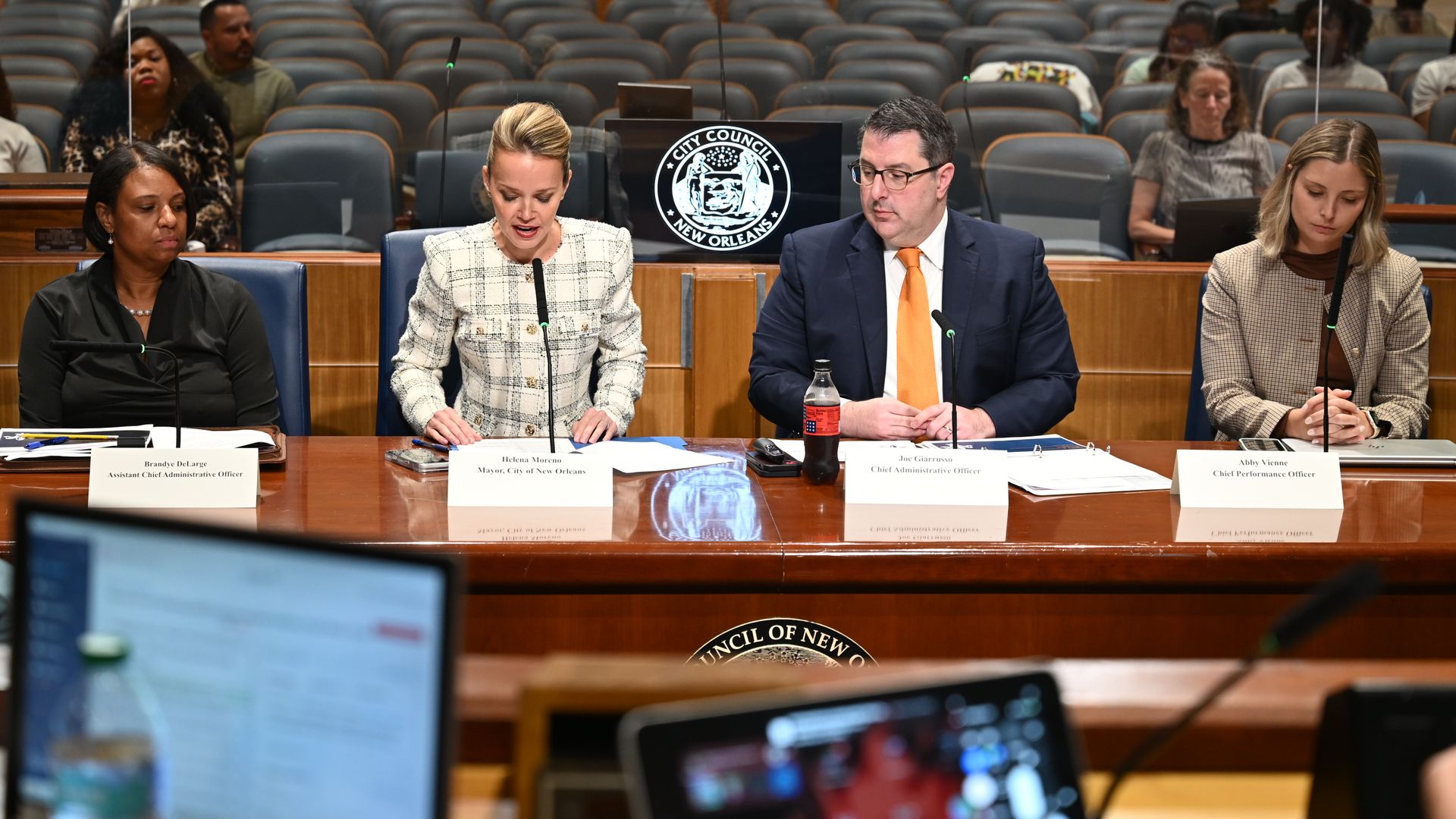 Three city officials sit at a council desk; middle woman reads from papers, man with an orange tie beside her. Behind them a circular seal reads "City Council New Orleans."