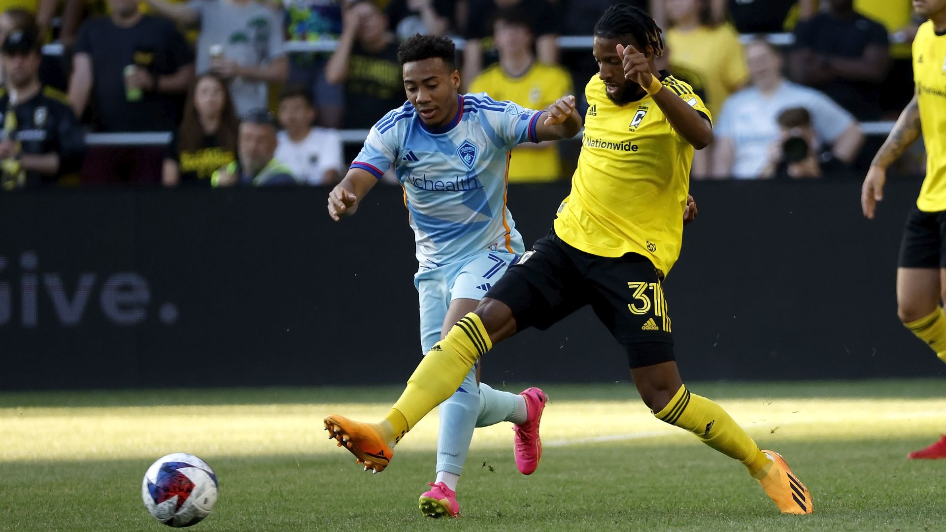 Two soccer players fight for a ball, with one extending his leg while another runs behind him, attempting to catch the ball.