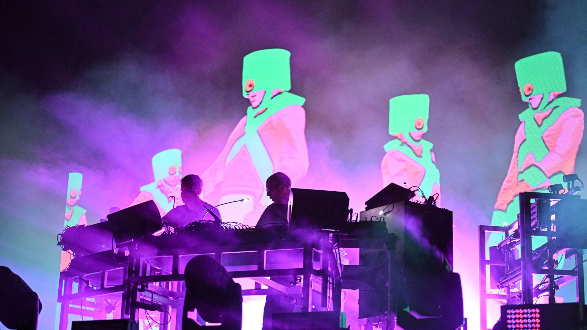 Photo of two men DJing on stage while graphics of people wearing masks appear on a projection screen behind them