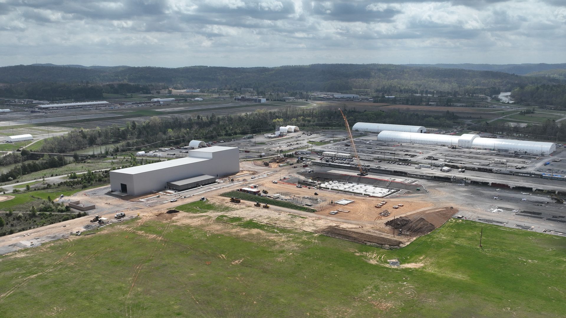 Aerial view of a construction site beside a gray warehouse and white hangars; a crane and equipment dot the dirt lots, with green fields and distant hills under a cloudy sky.