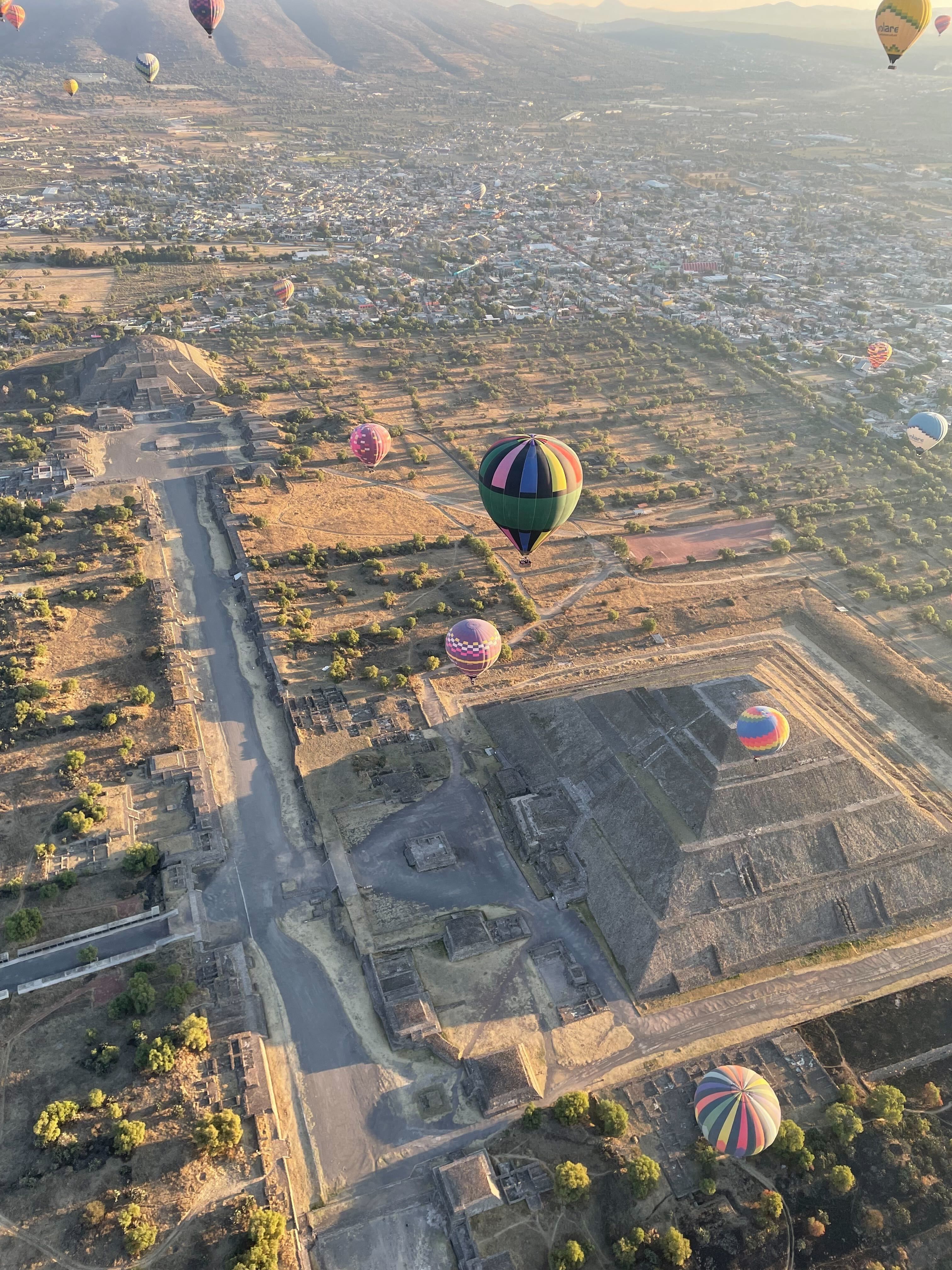 An aerial view of a desert archaeological site with stepped pyramids and ruins, a road, and a town in the distance, while many colorful hot air balloons float above at sunrise.