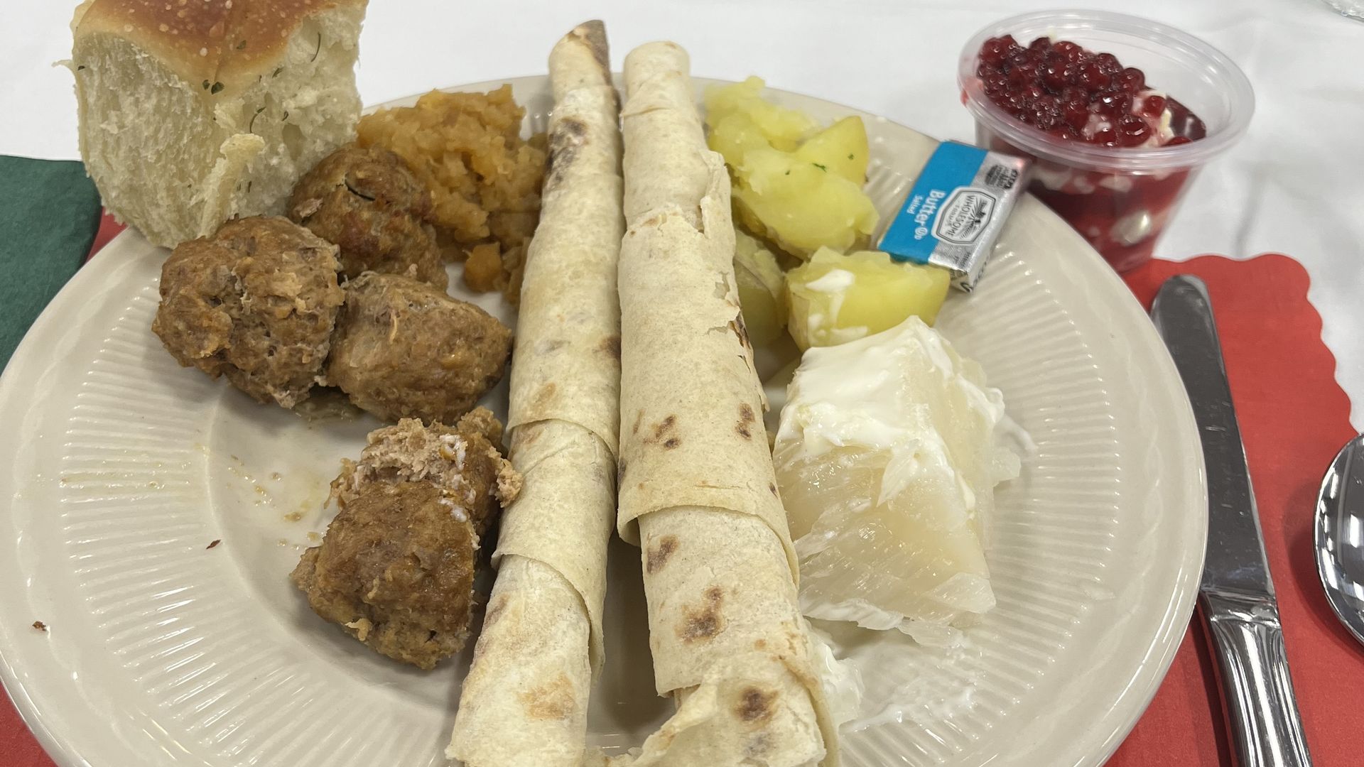 Plate with Swedish food including meatballs, lingonberries in a cup, boiled potatoes, flatbread, bread roll, and white sauce on Lutefisk, all on a table with red and green placemats.